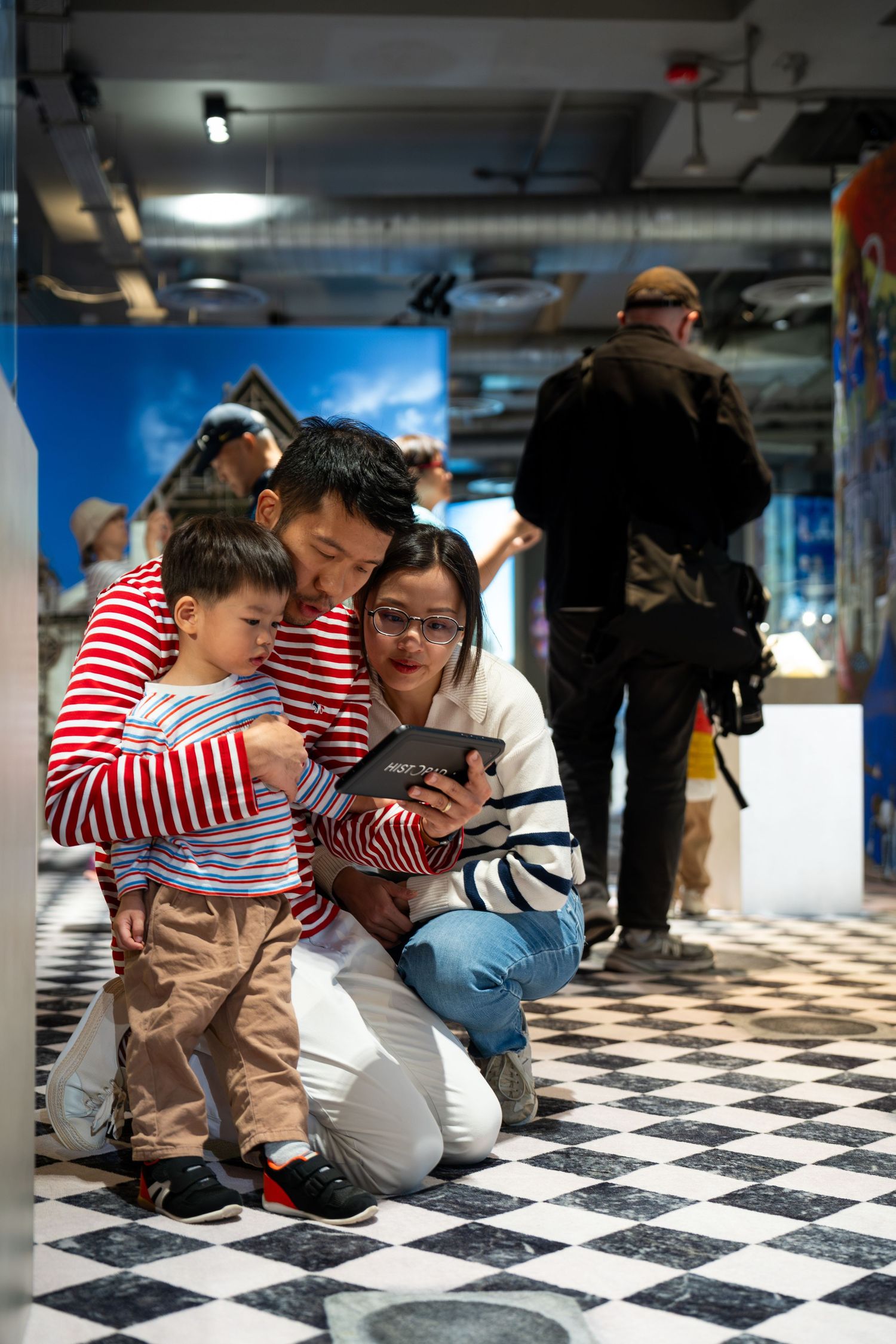 Asian family with young child views tablet together at colorful interactive museum exhibit.