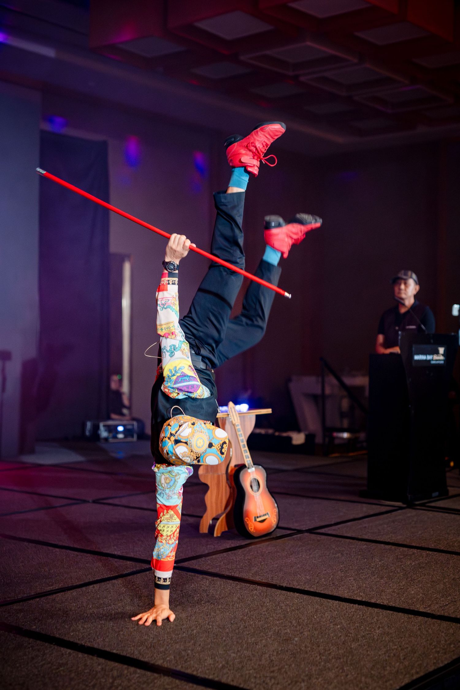 Acrobat performing a one-handed handstand while holding a red pole at an indoor event with colorful stage lighting.