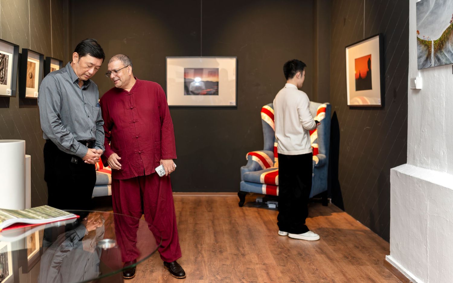Two men examine artwork in a gallery space while another person browses framed photos on the wall.
