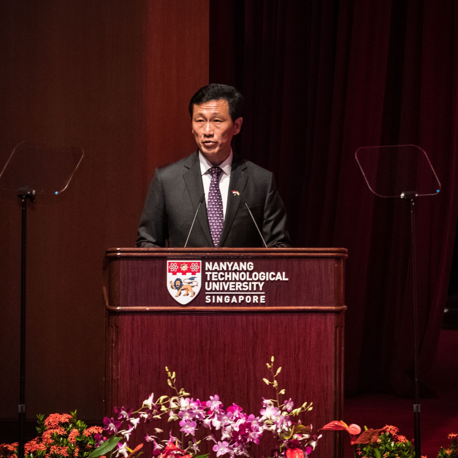 Speaker at Nanyang Technological University Singapore podium, wearing suit and purple tie, with floral arrangement.