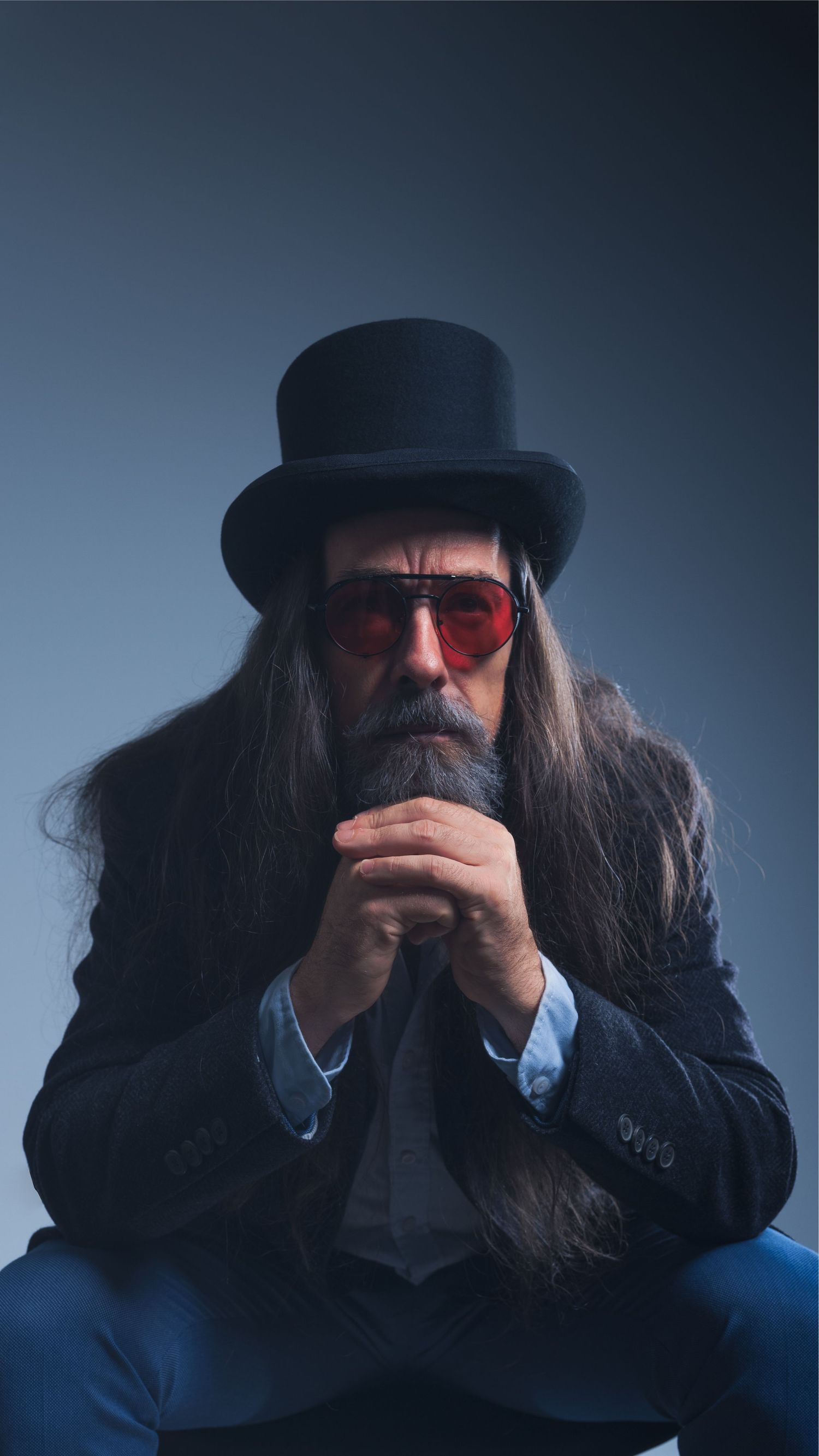 Bearded man with long hair wearing a black top hat and red tinted sunglasses, sitting in a dramatic studio portrait.