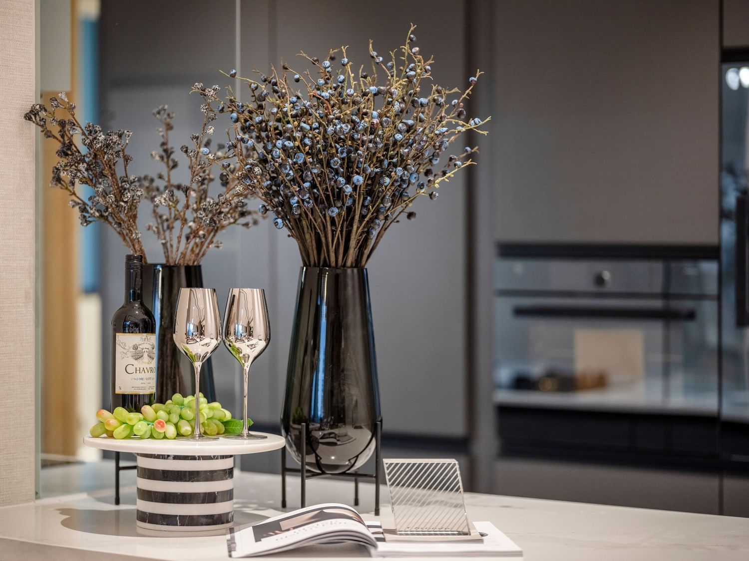 Modern kitchen counter with dark vases of dried flowers, wine glasses, grapes, and a wine bottle in an elegant setting.
