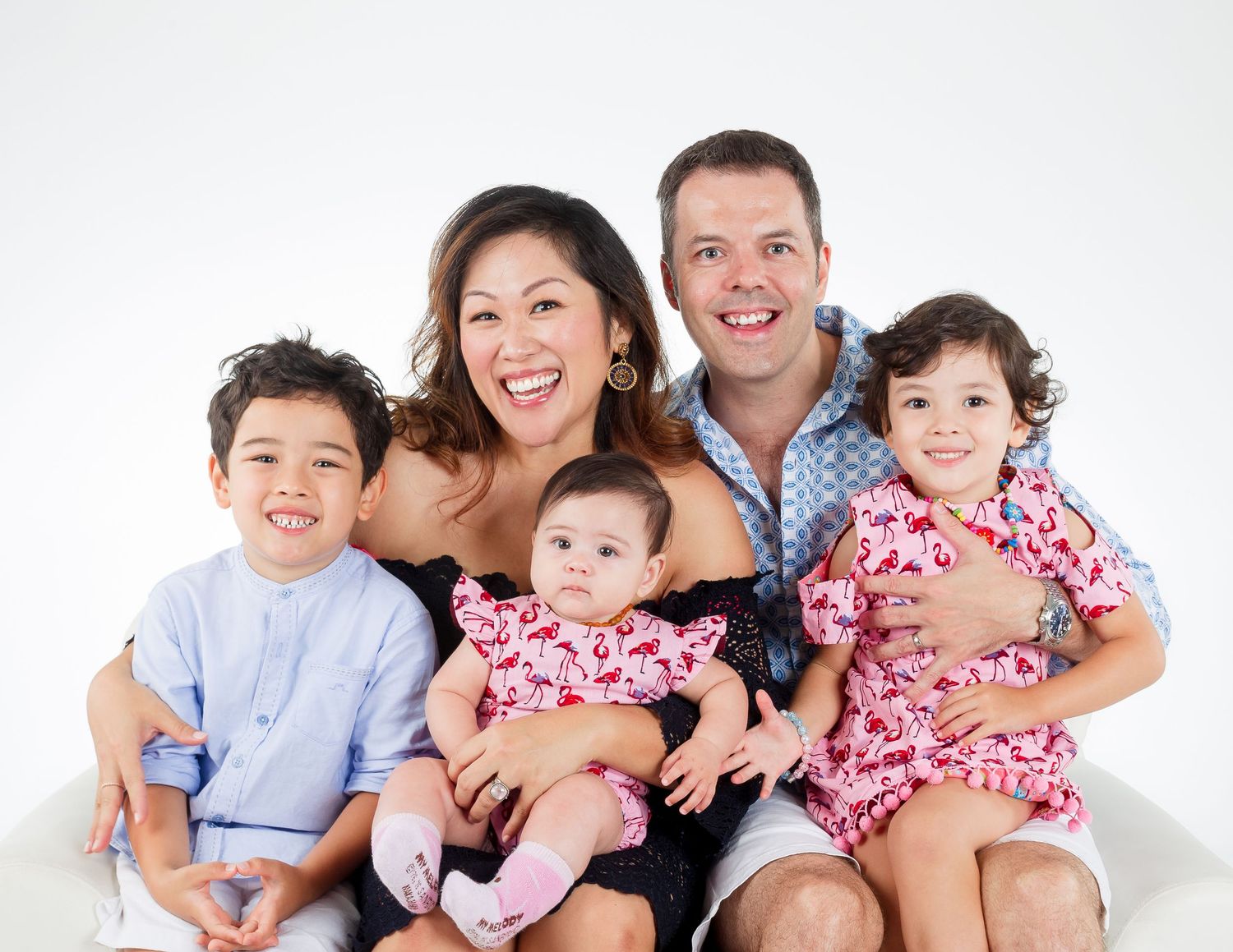 Cheerful family of five with three young children smiles together in a bright white studio portrait.