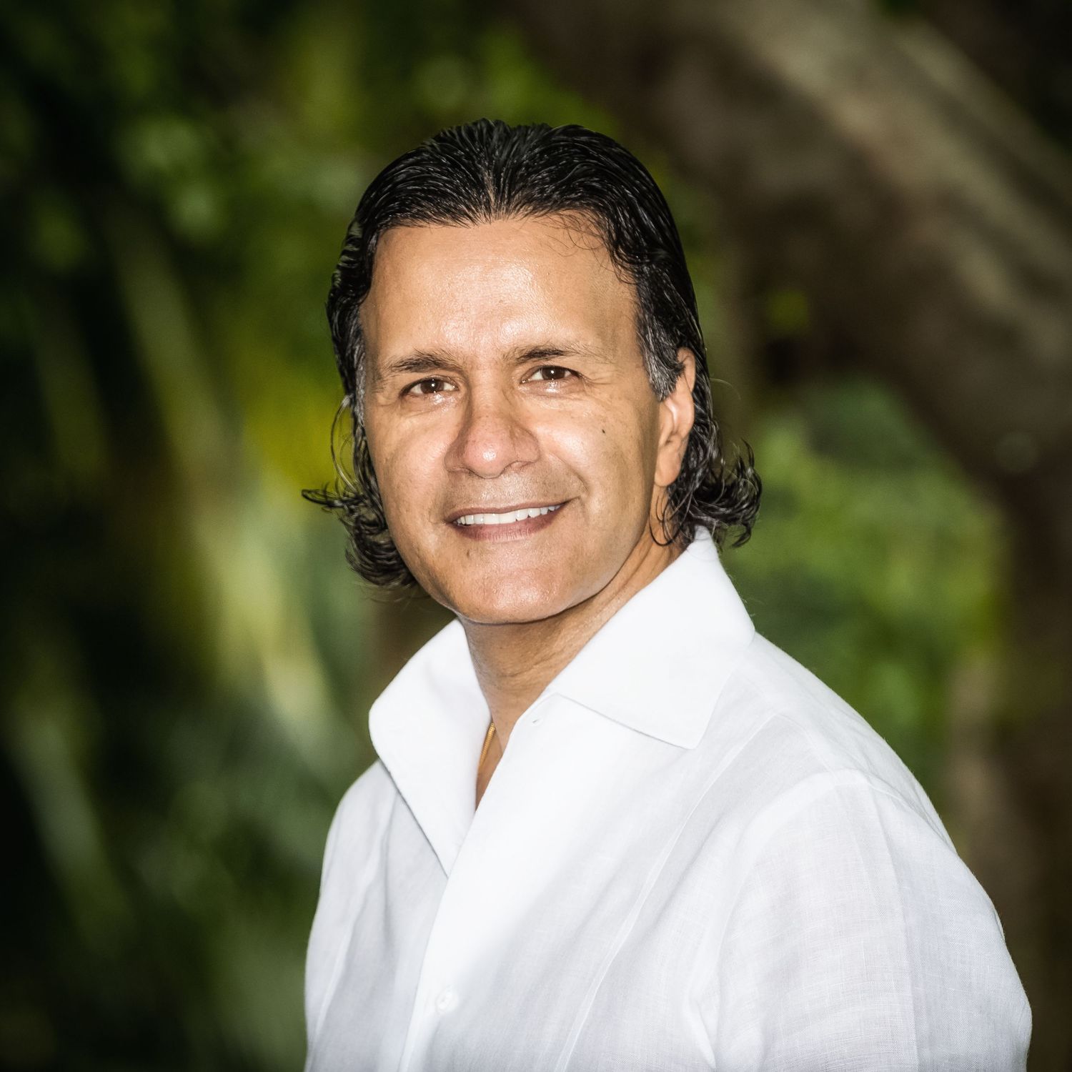 Smiling middle-aged man with dark hair poses outdoors in a white shirt among lush green foliage.
