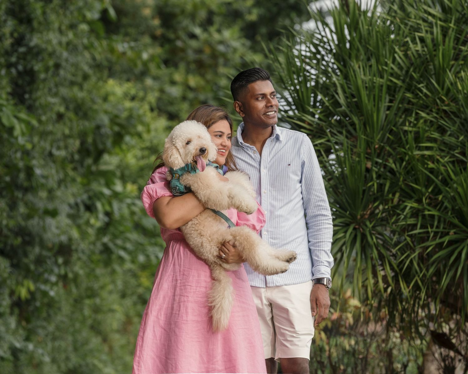 Couple holding a fluffy white dog outdoors surrounded by lush green tropical foliage.