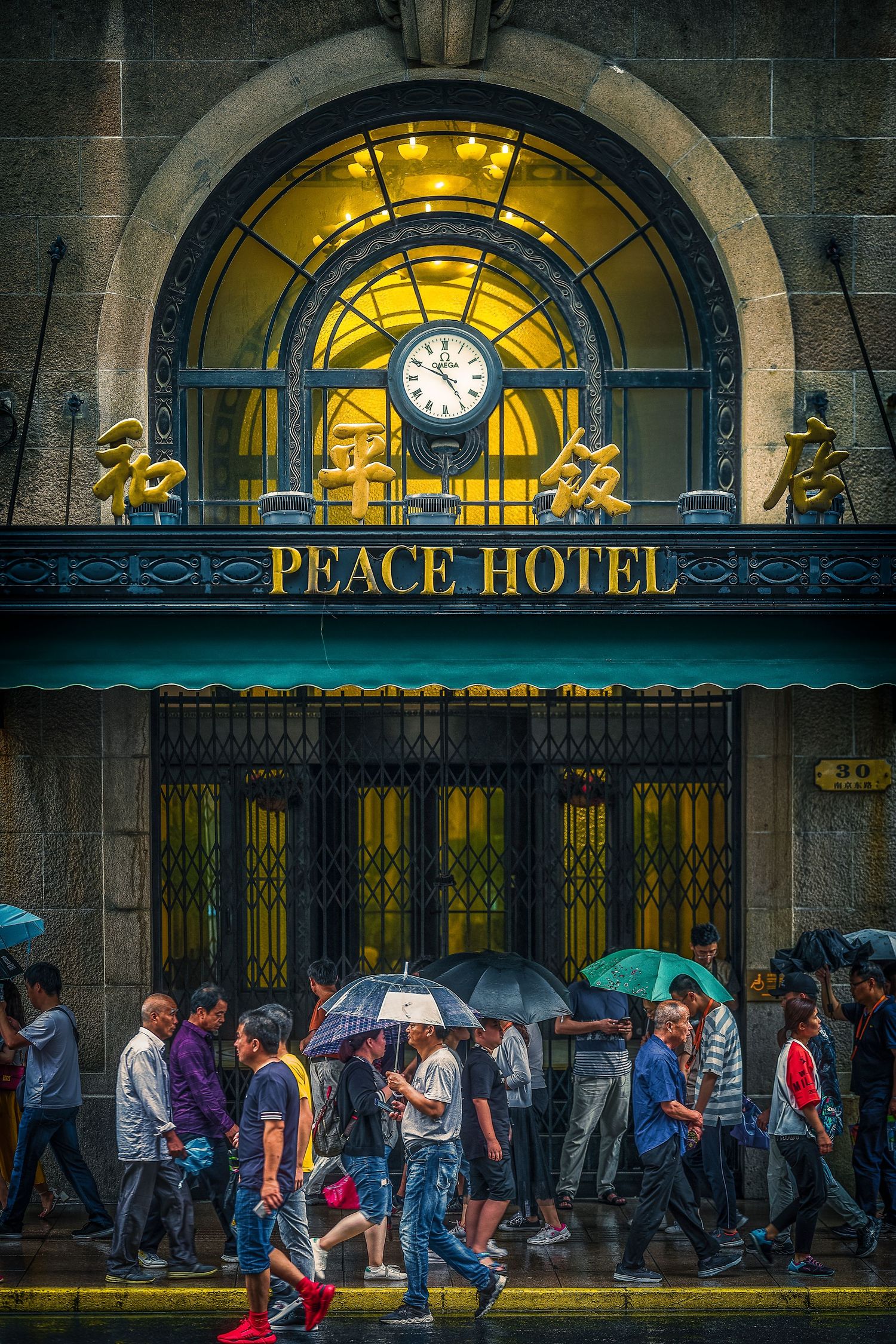 Crowds with umbrellas walk past the historic Peace Hotel entrance in Shanghai, China, on a rainy day.