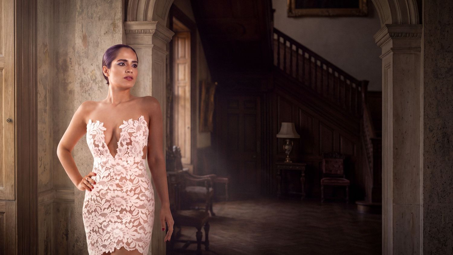 Elegant woman in a fitted lace strapless gown poses in a grand historic mansion with staircase.