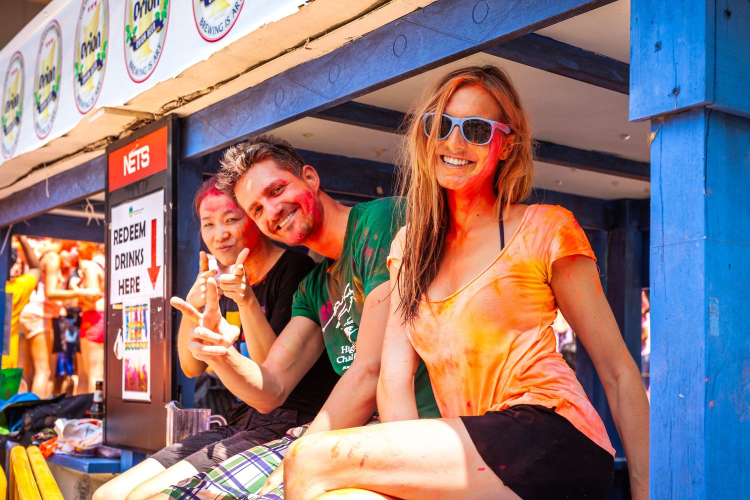 Three friends covered in orange Holi powder smile at a festive outdoor event booth.