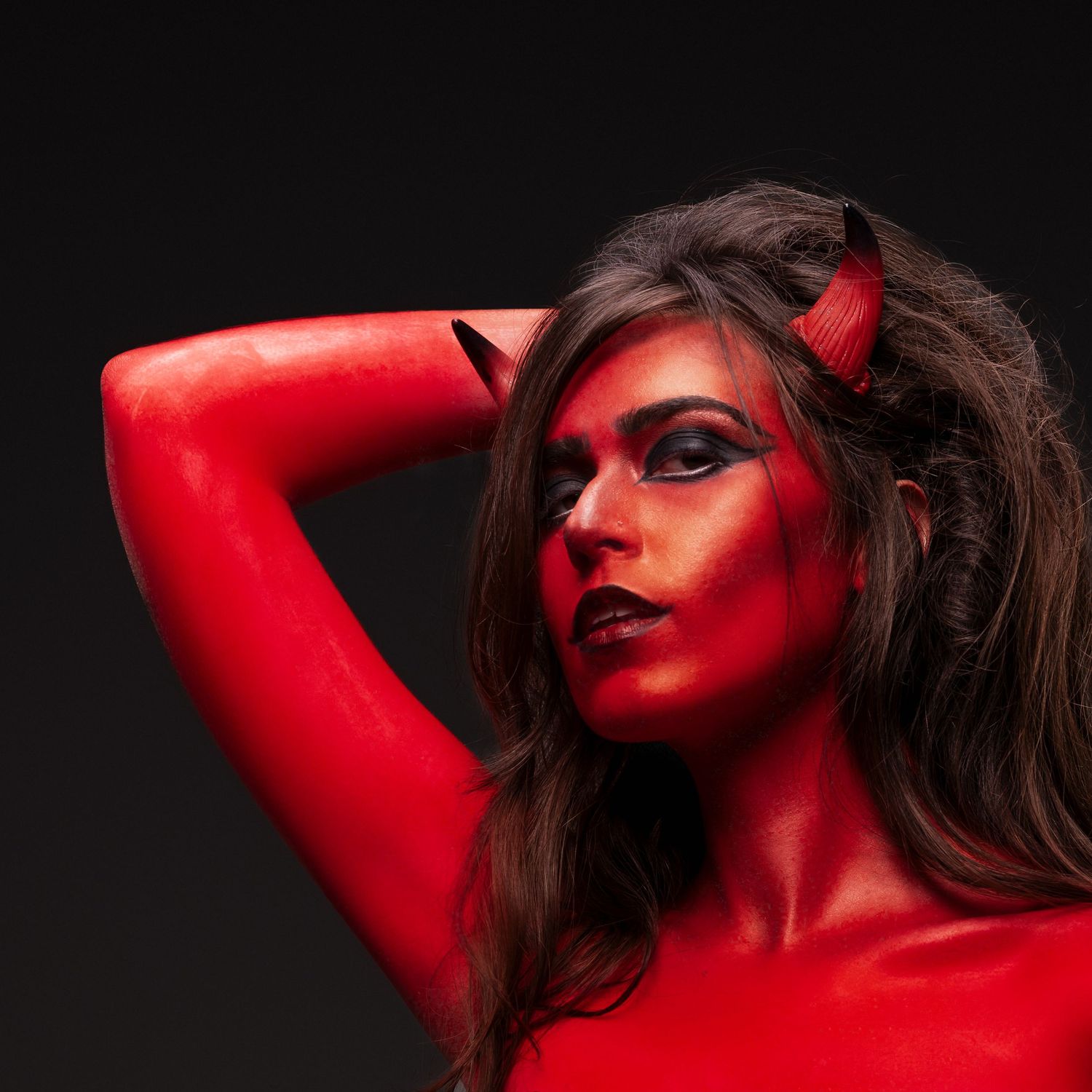Woman in red body paint and devil horns costume poses dramatically against dark background.