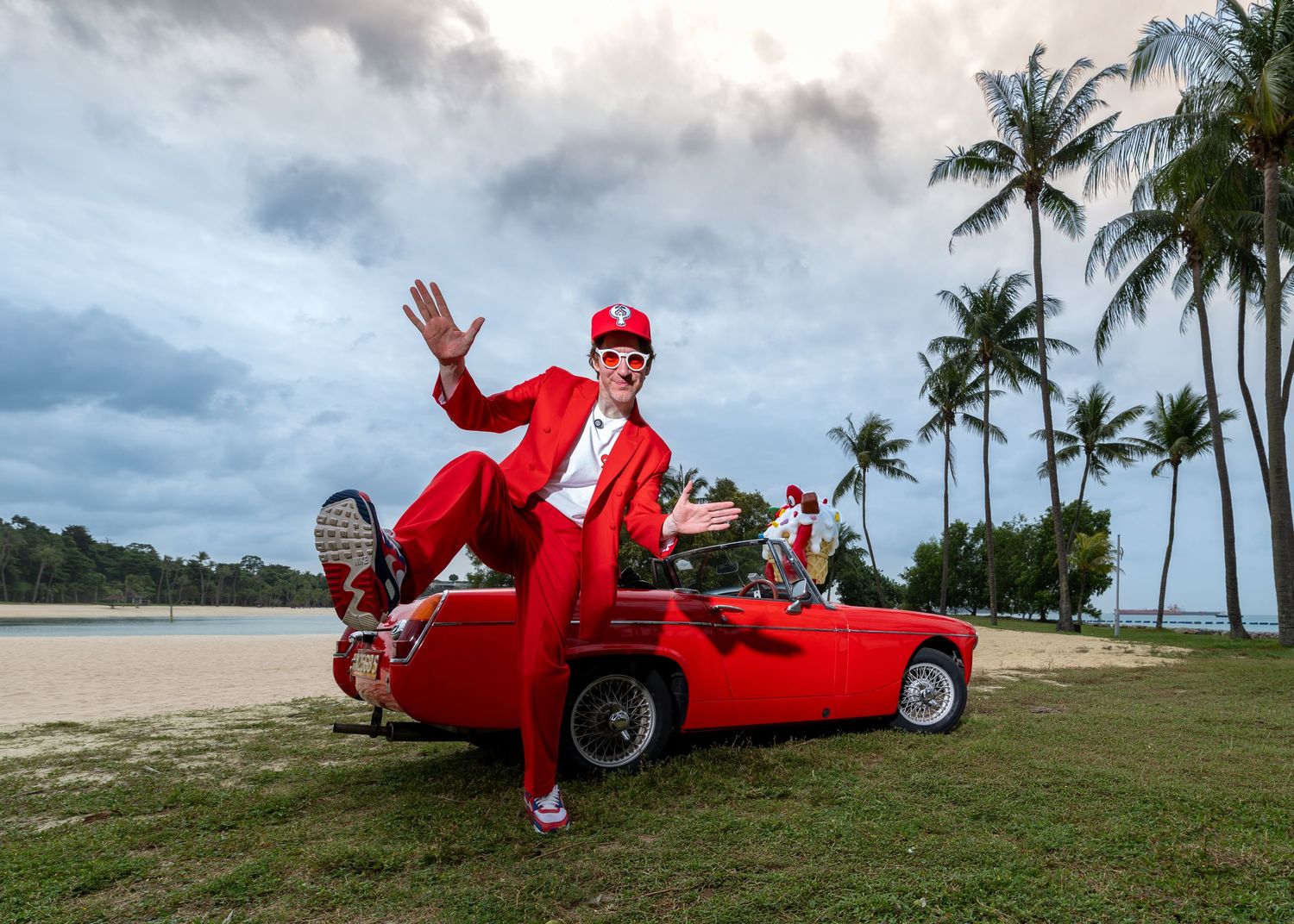 Man in red suit poses on red vintage car near palm trees and beach on cloudy day.