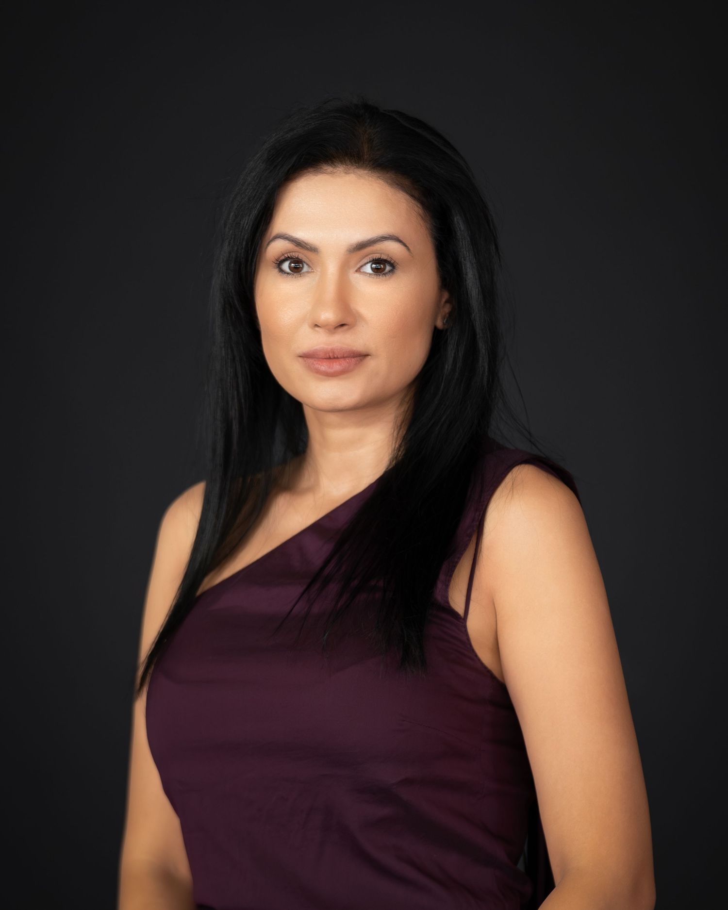 Professional headshot of a woman with long black hair wearing a dark burgundy one-shoulder top against a dark background.