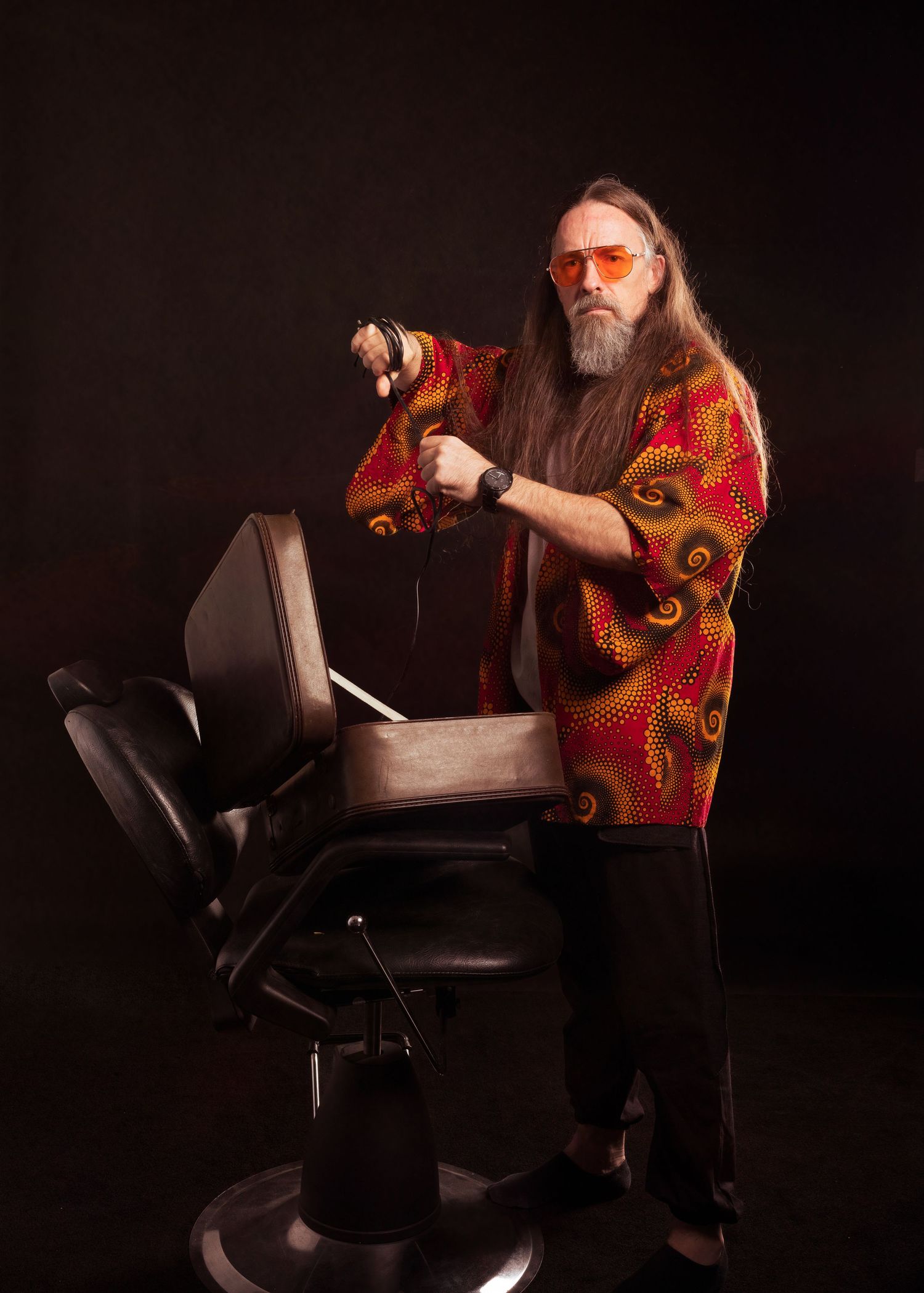Long-haired bearded man in colorful red shirt and orange sunglasses holding scissors near a barber chair.