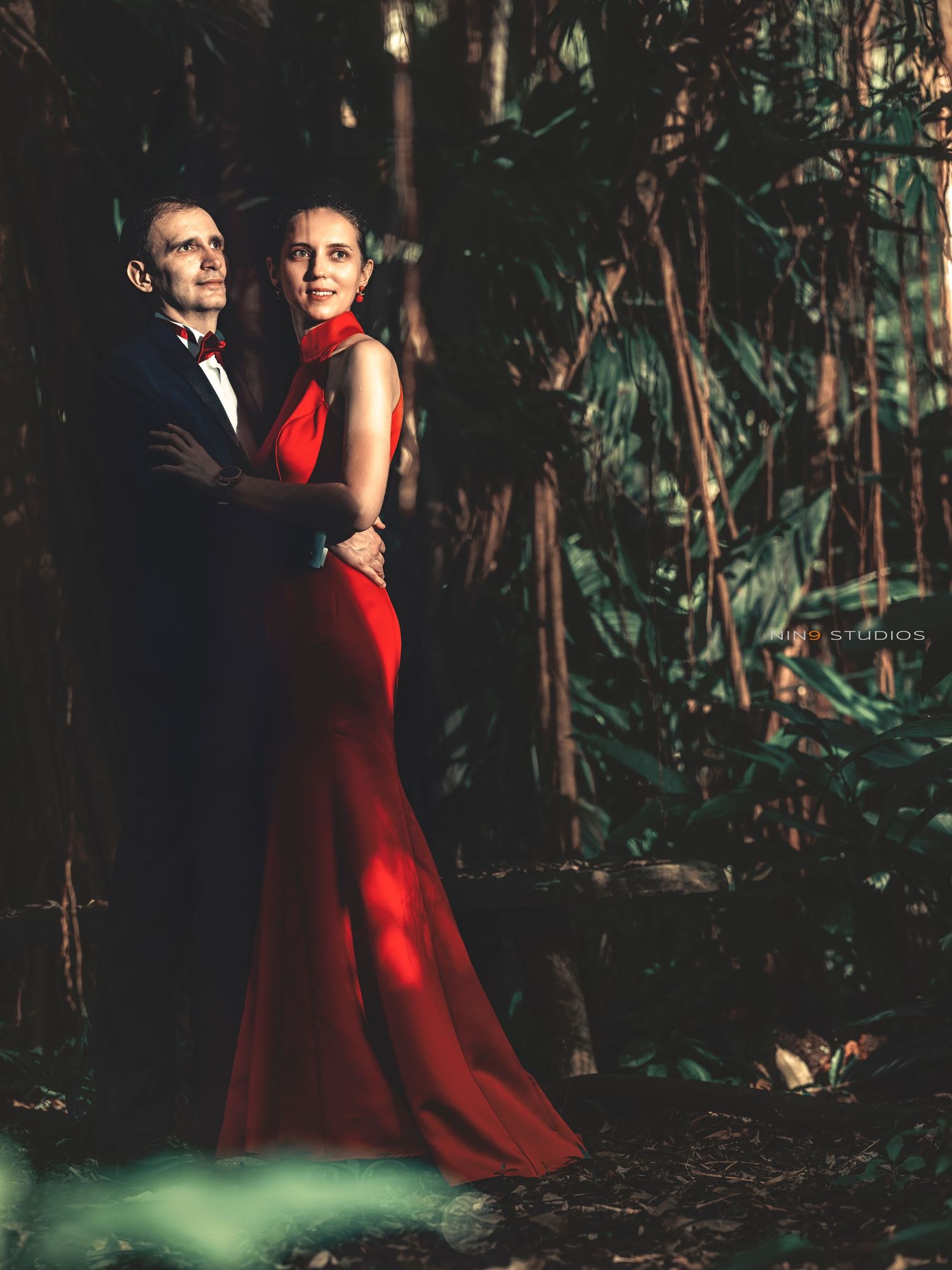 Couple in elegant formalwear posing in lush forest, woman in red gown, man in black tuxedo.