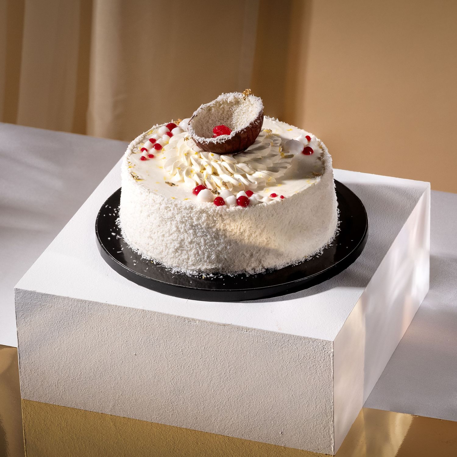 White coconut cake topped with shredded coconut, red berry decorations, and a halved coconut on a black cake board.
