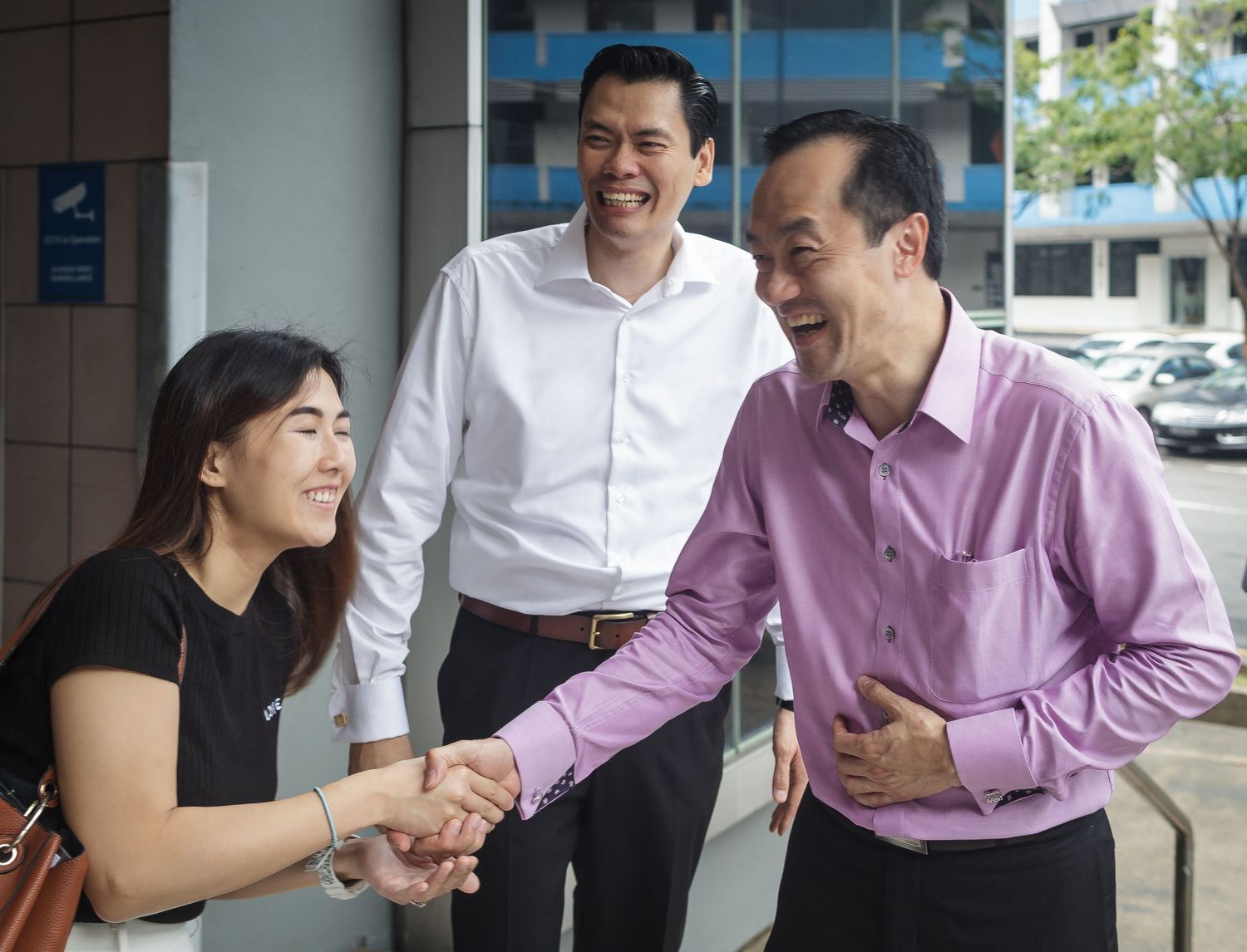 Two men and a young woman share a warm handshake greeting outside a building in Singapore.