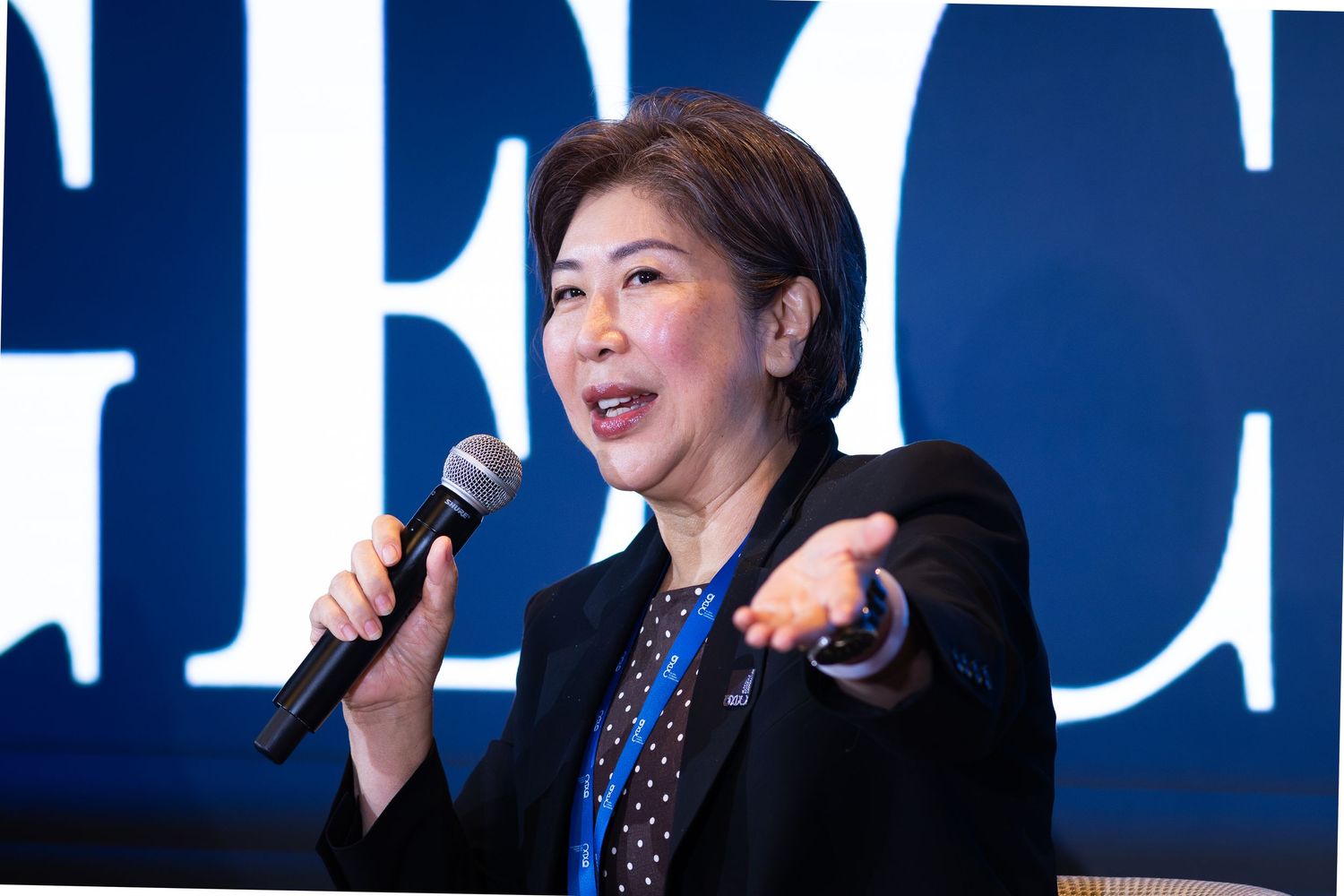A woman in a black blazer gestures while speaking at a professional conference with a blue backdrop.