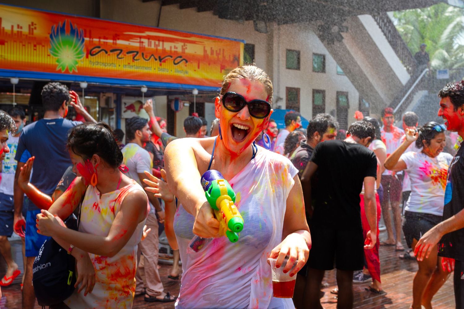 Woman with sunglasses joyfully playing with water gun at colorful Holi festival celebration outdoors.