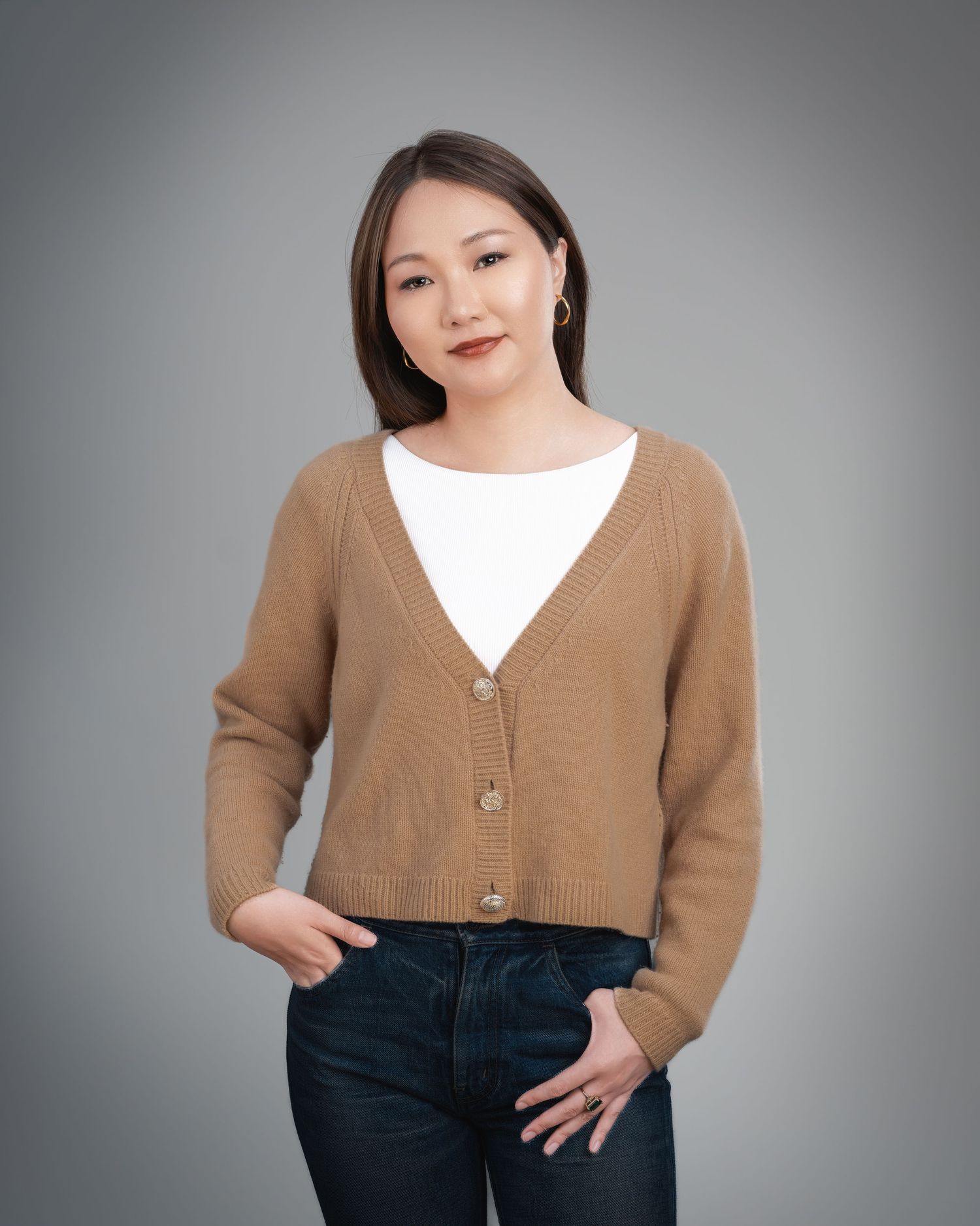 Young woman in tan cardigan and jeans posing with hands in pockets on gray background.