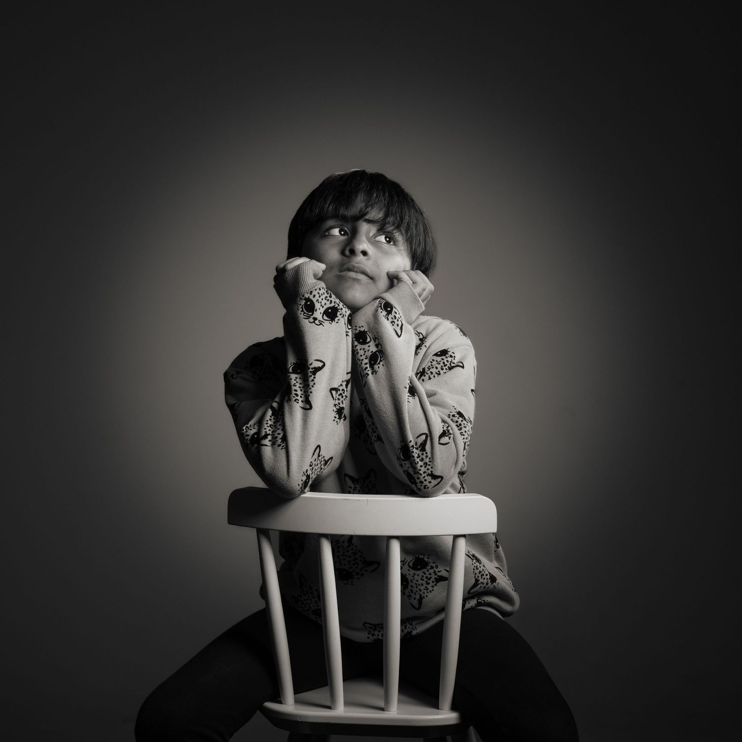 Black and white portrait of a pensive child sitting on a white wooden chair, resting face in hands, looking upward.