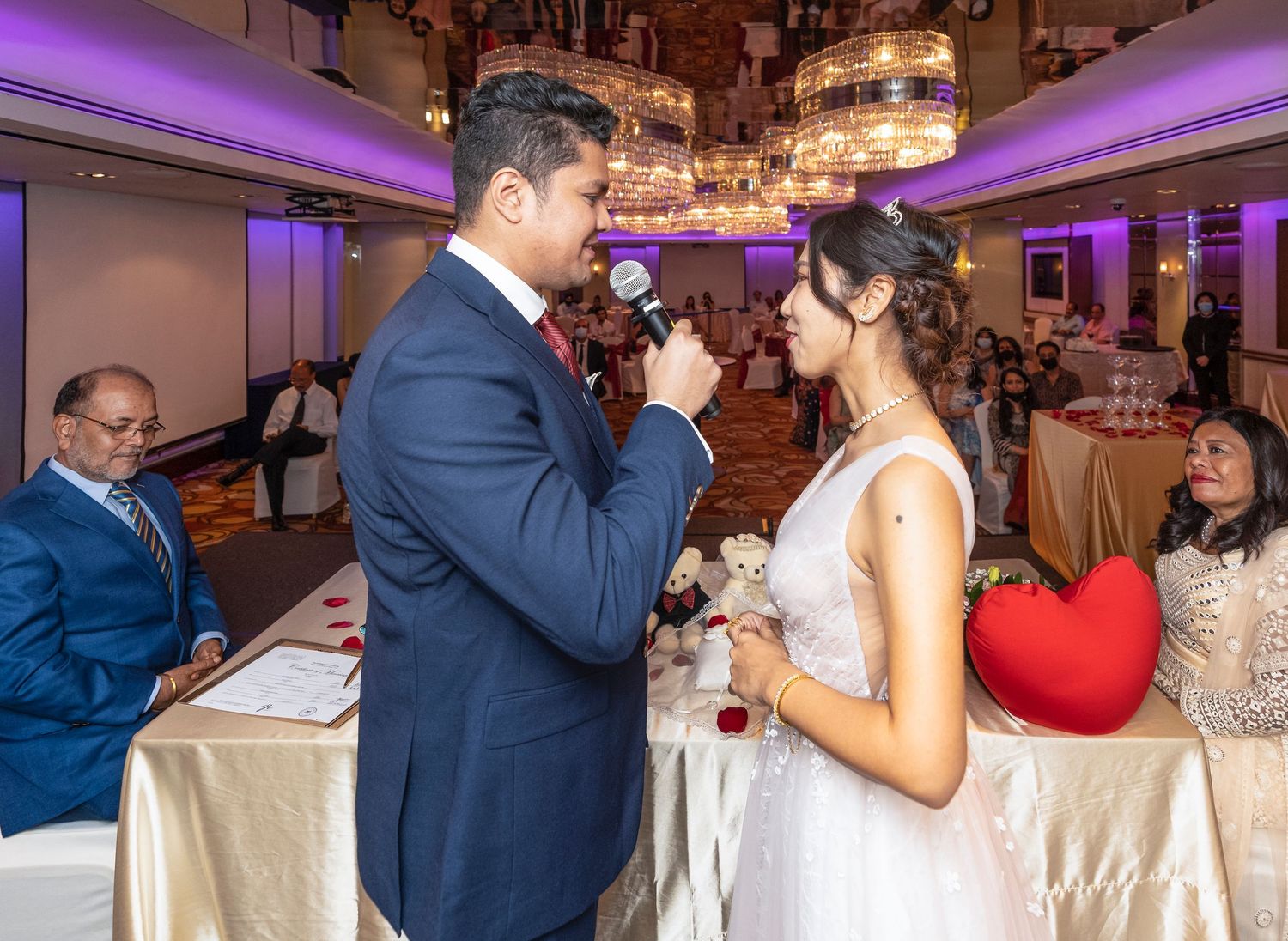 Groom speaks into microphone during elegant wedding reception as bride listens in decorated ballroom.