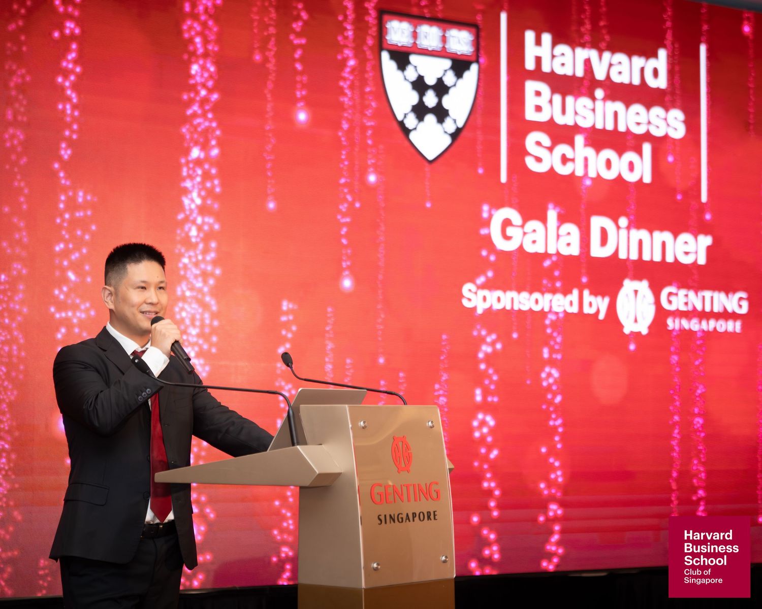 Man speaking at Harvard Business School Gala Dinner podium, sponsored by Genting Singapore, at a red-themed event.