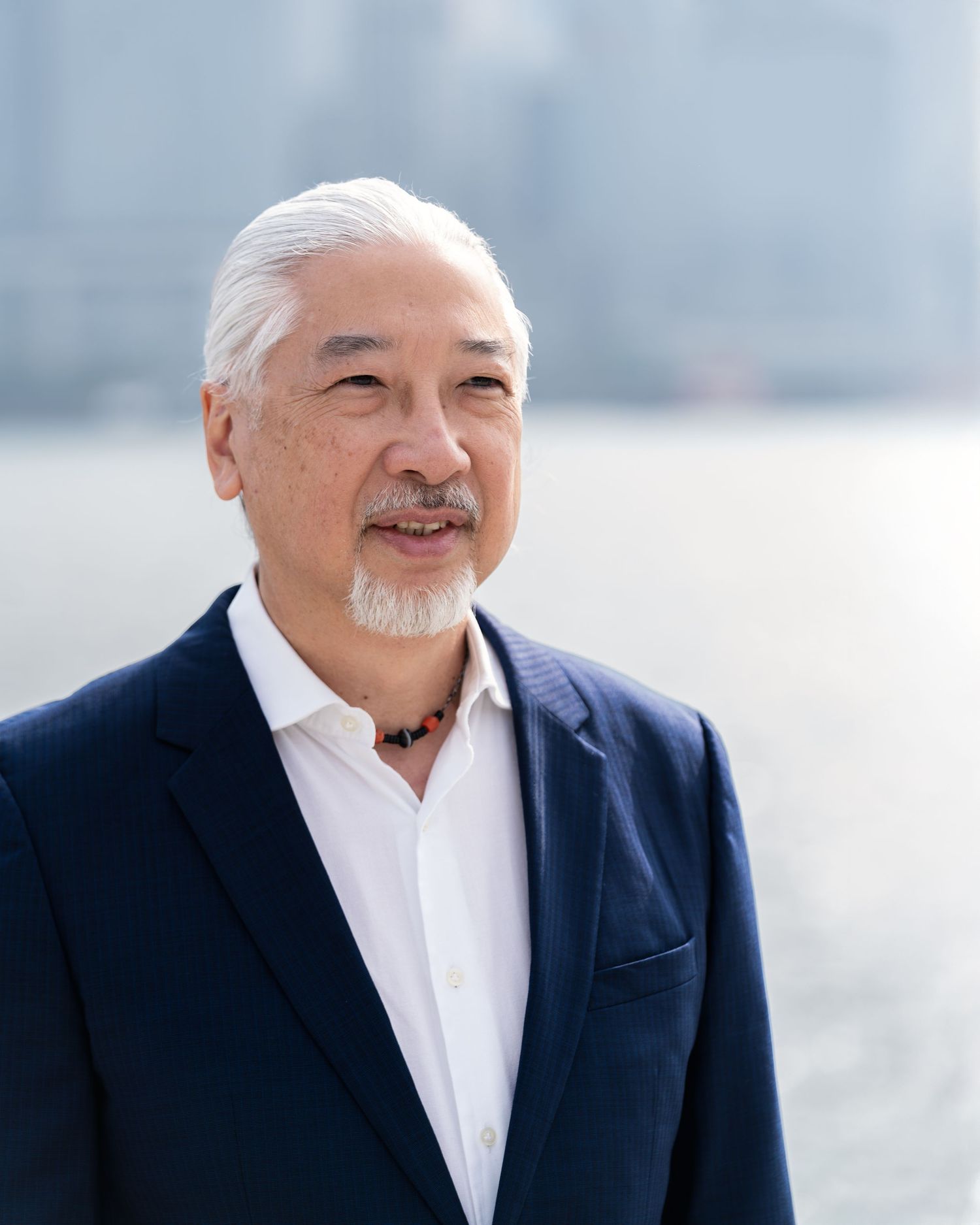 An older man with white hair and a goatee wearing a navy blazer outdoors near a waterfront.