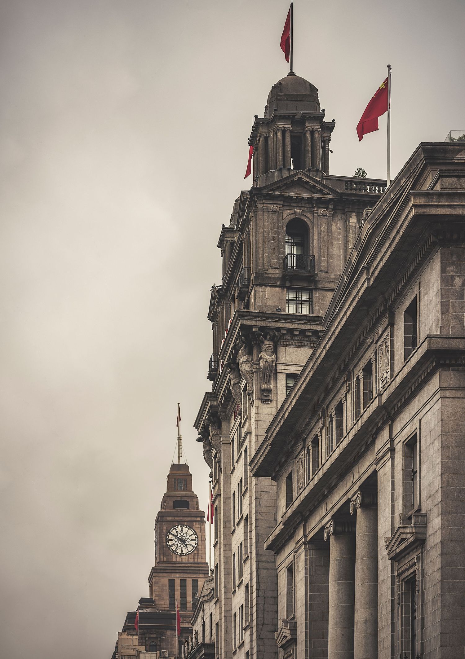 Historic colonial architecture with red flags and clock tower on the Bund in Shanghai under overcast skies.