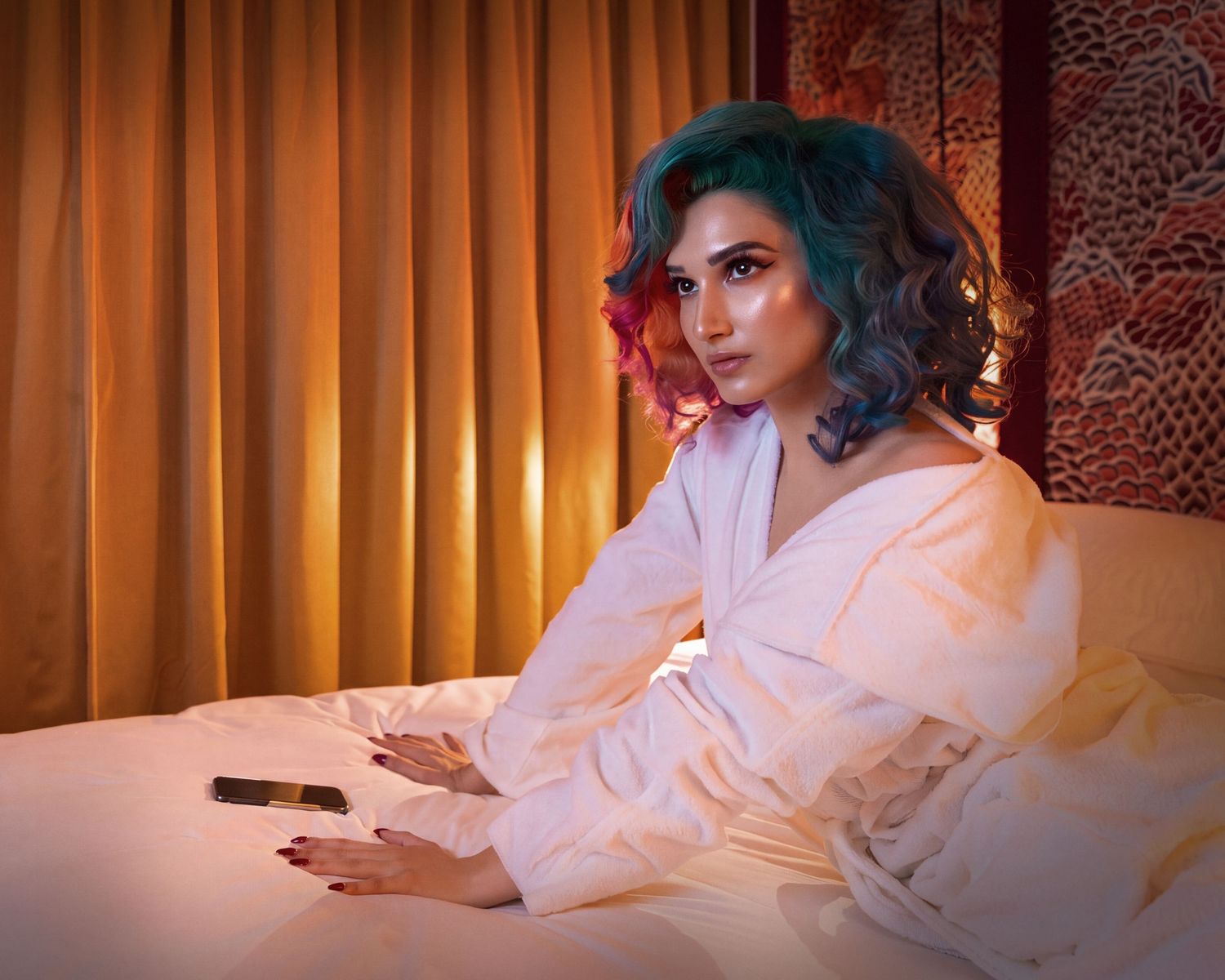 Woman with colorful teal and pink hair in white robe sitting on bed with warm golden curtain backdrop.