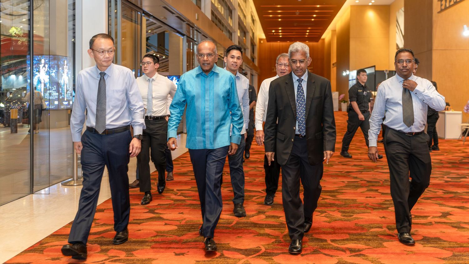 Group of businessmen in formal attire walk confidently through a carpeted conference hall corridor.