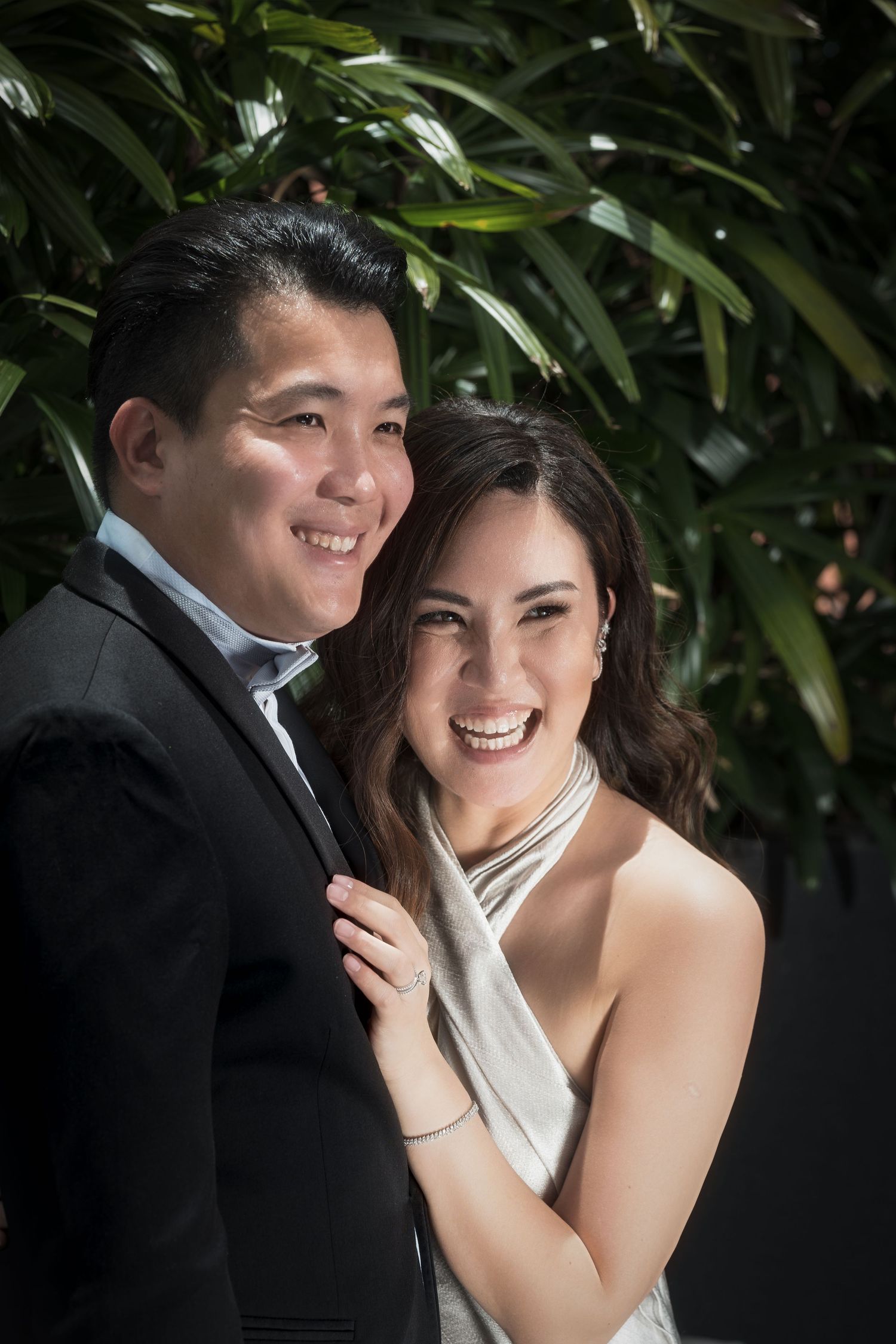 Smiling Asian couple posing together outdoors, man in black suit and woman in elegant white halter dress, with lush green foliage.