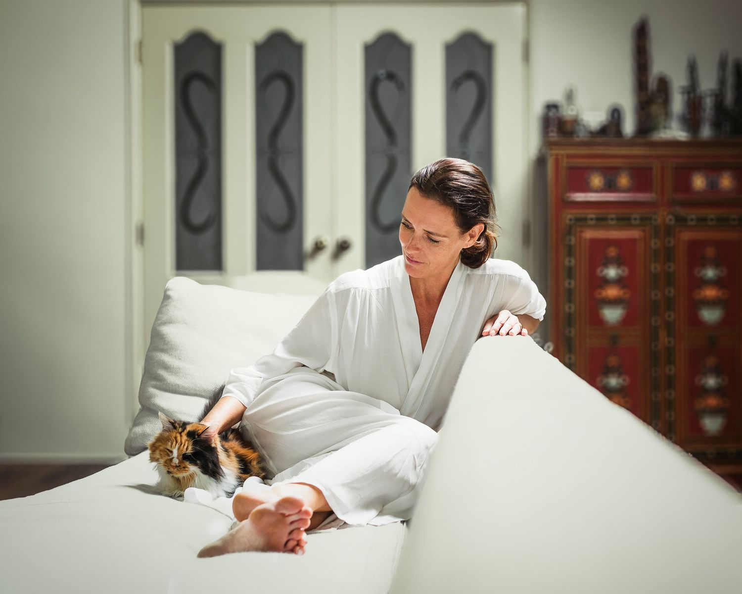 Woman in white robe relaxing on sofa while petting a tabby cat in a stylish living room.