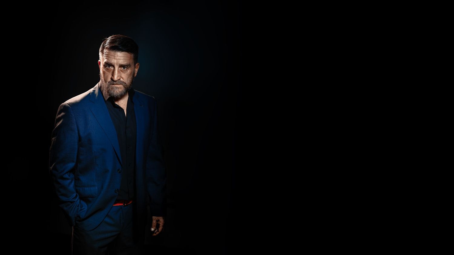 Confident man in blue suit posing against dark background with dramatic lighting.