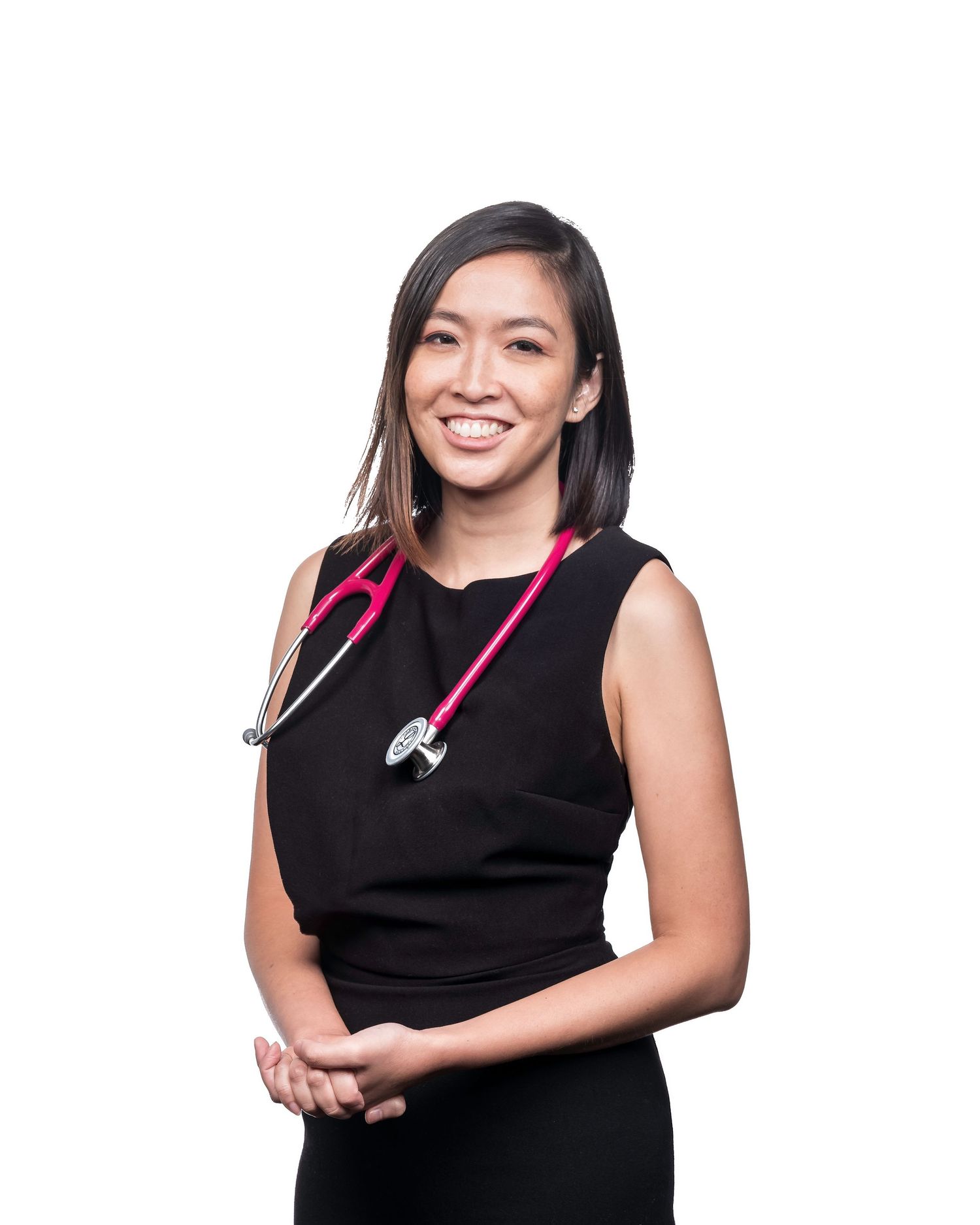 Smiling young female doctor in black dress with pink stethoscope poses against a white background.