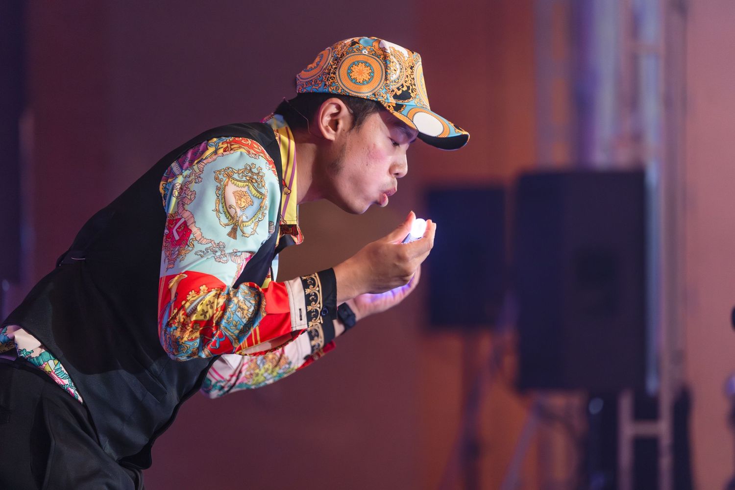 Performer in colorful patterned jacket and cap leaning forward on stage under dramatic lighting.
