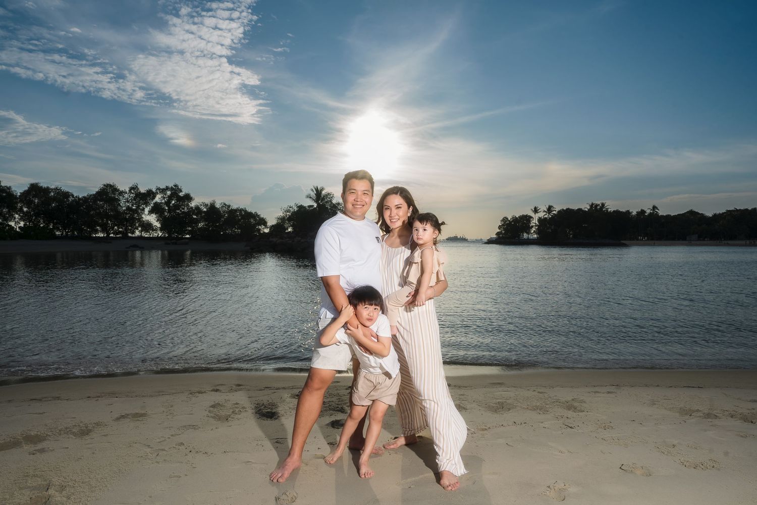 Family of four posing on a sandy beach at sunset with calm water and lush trees in the background.