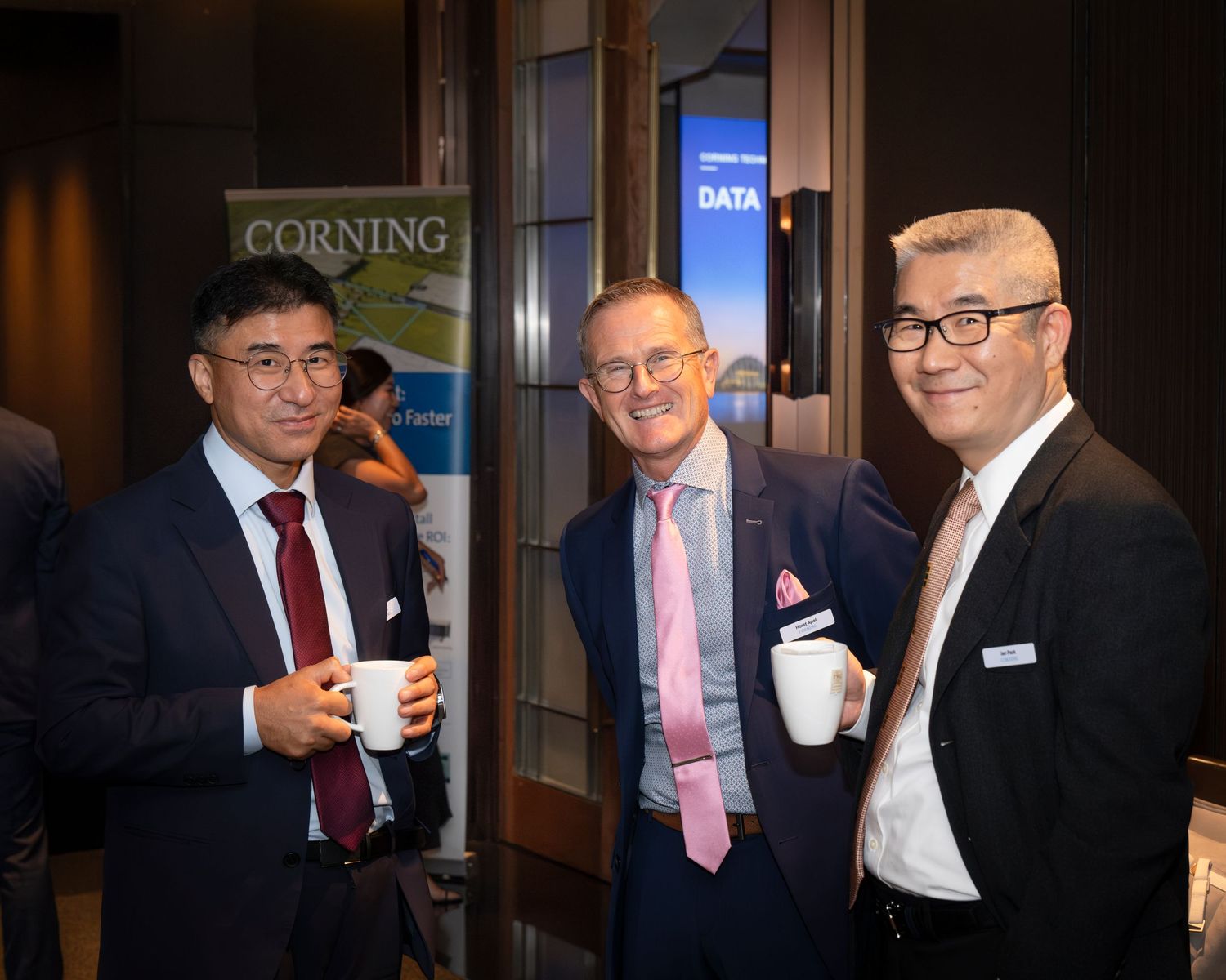 Three businessmen networking at a Corning corporate event, smiling and holding coffee cups in a modern venue.