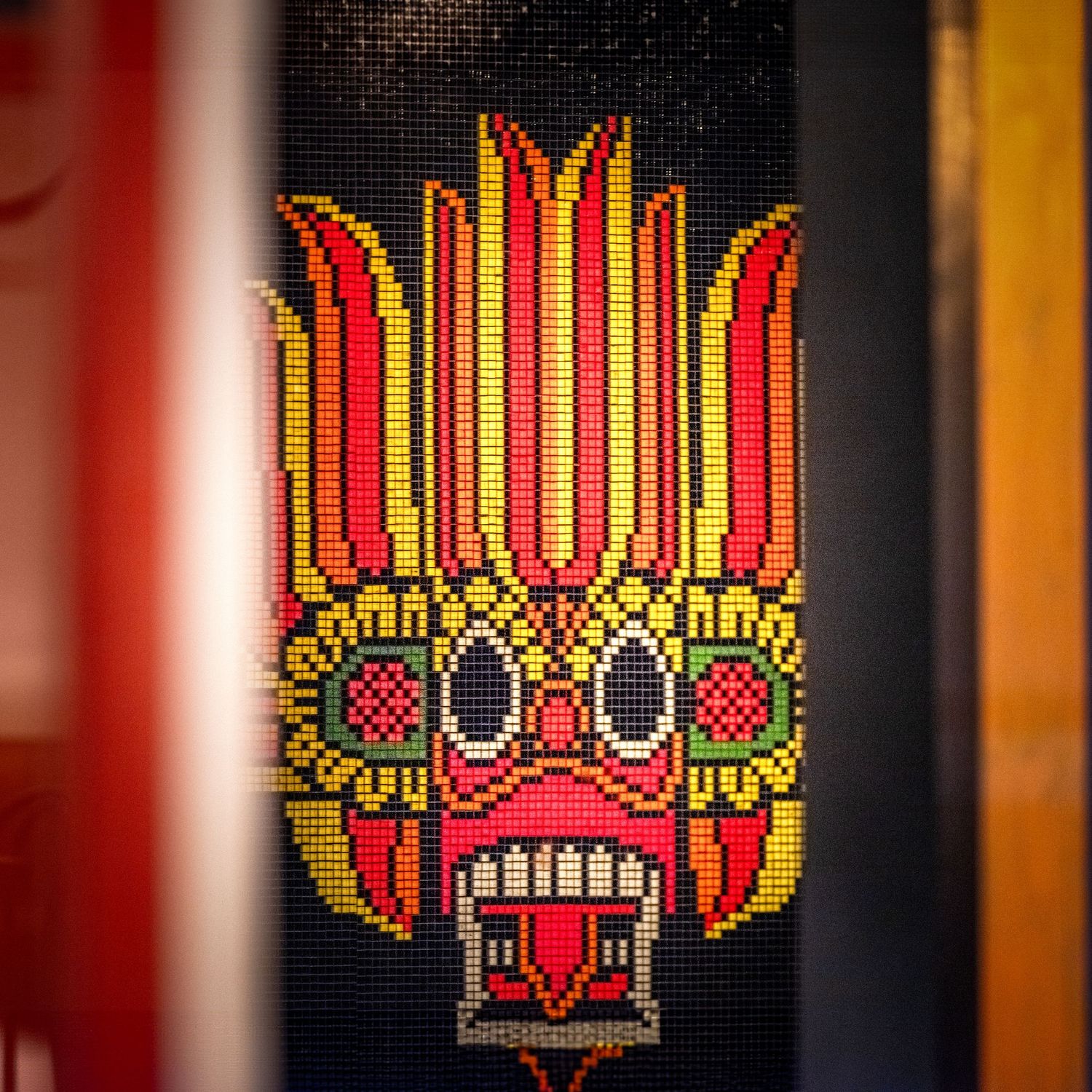 Colorful pixel art Balinese demon mask with red, yellow, and green mosaic tiles on dark background.