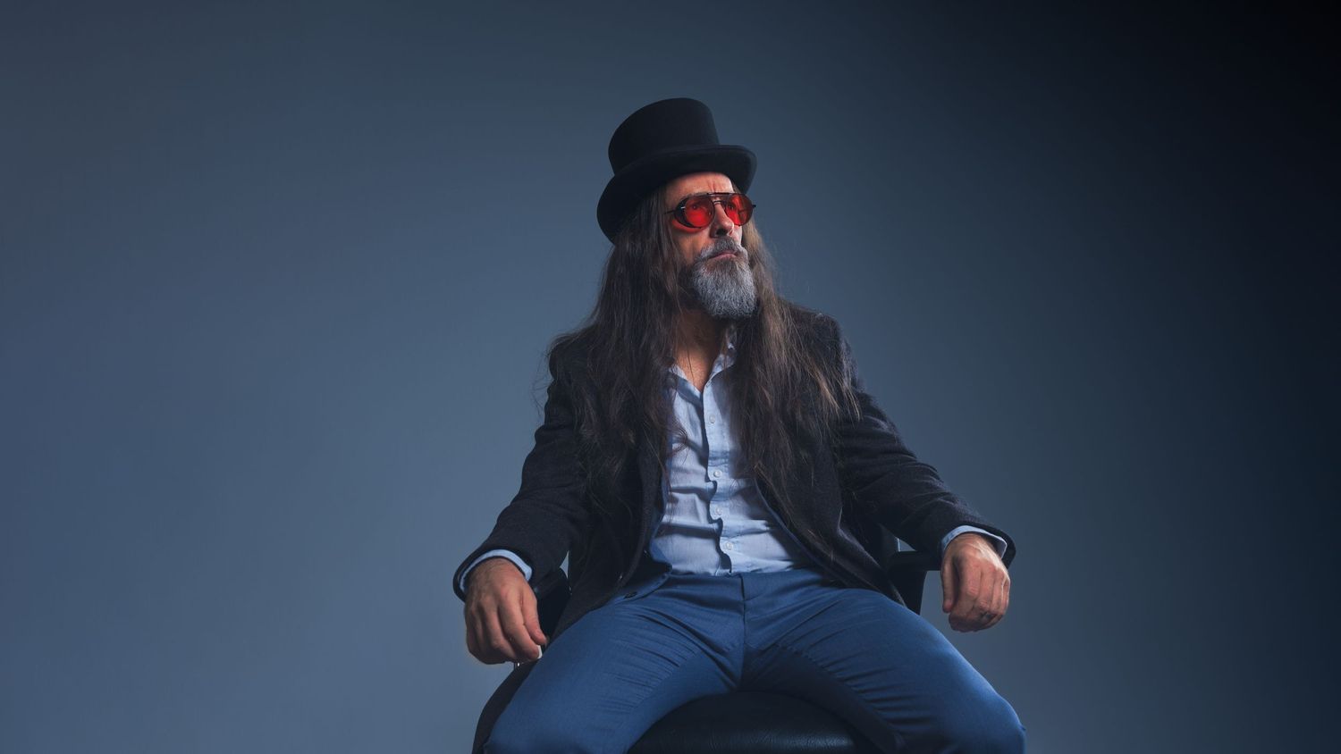 Man with long hair wearing a black top hat, red sunglasses, and blazer seated against a gray background.