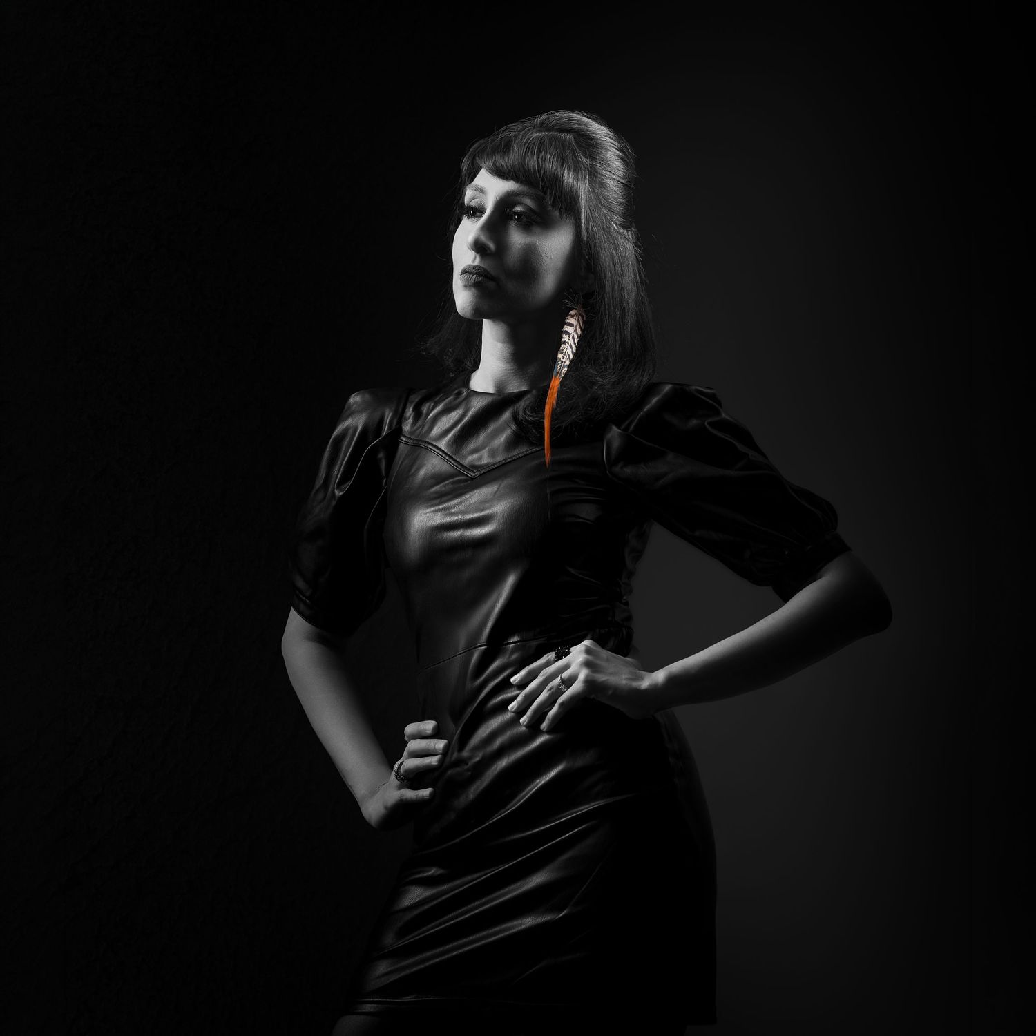 Woman in black leather dress poses dramatically against dark background, holding a colorful feather.