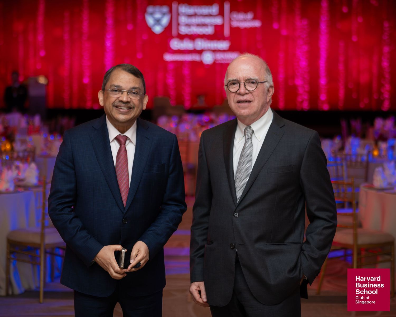 Two men in suits pose at Harvard Business School Gala Dinner event with red-lit venue backdrop.