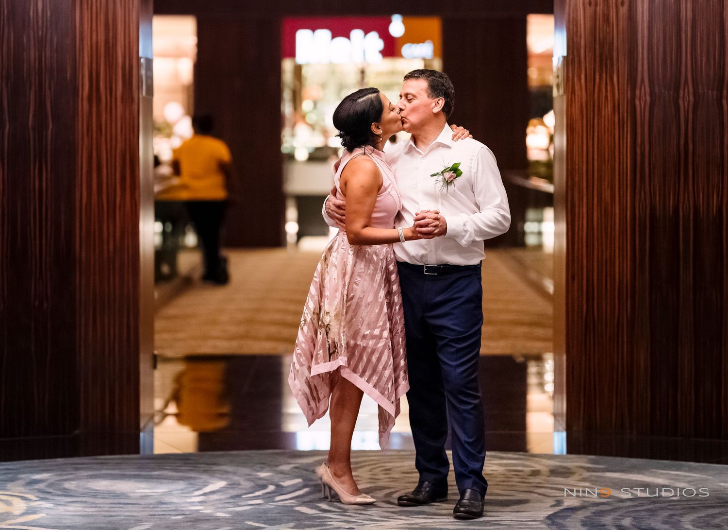 Couple sharing a romantic kiss in an elegant hotel lobby, woman in pink dress, man in navy suit with boutonniere.