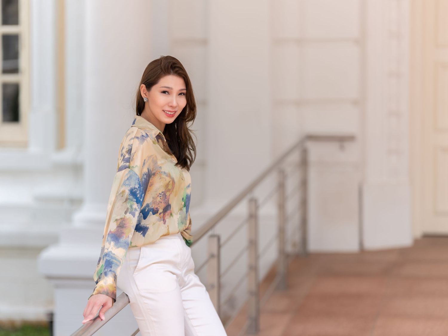 Young woman in floral blouse and white pants posing outdoors near building steps, smiling over shoulder.