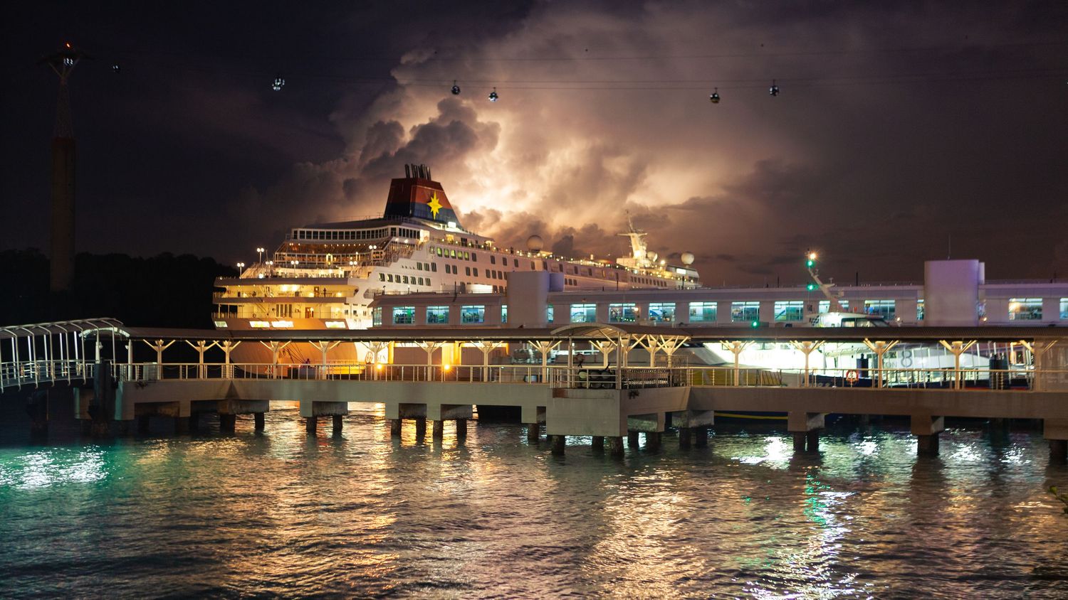 Large cruise ship docked at illuminated pier at night with dramatic stormy sky reflected in calm harbor waters.