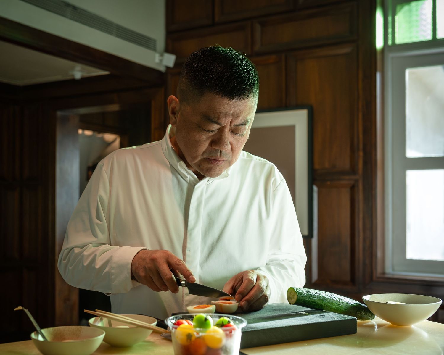 A chef in white uniform carefully cuts vegetables on a wooden cutting board in a traditional kitchen.