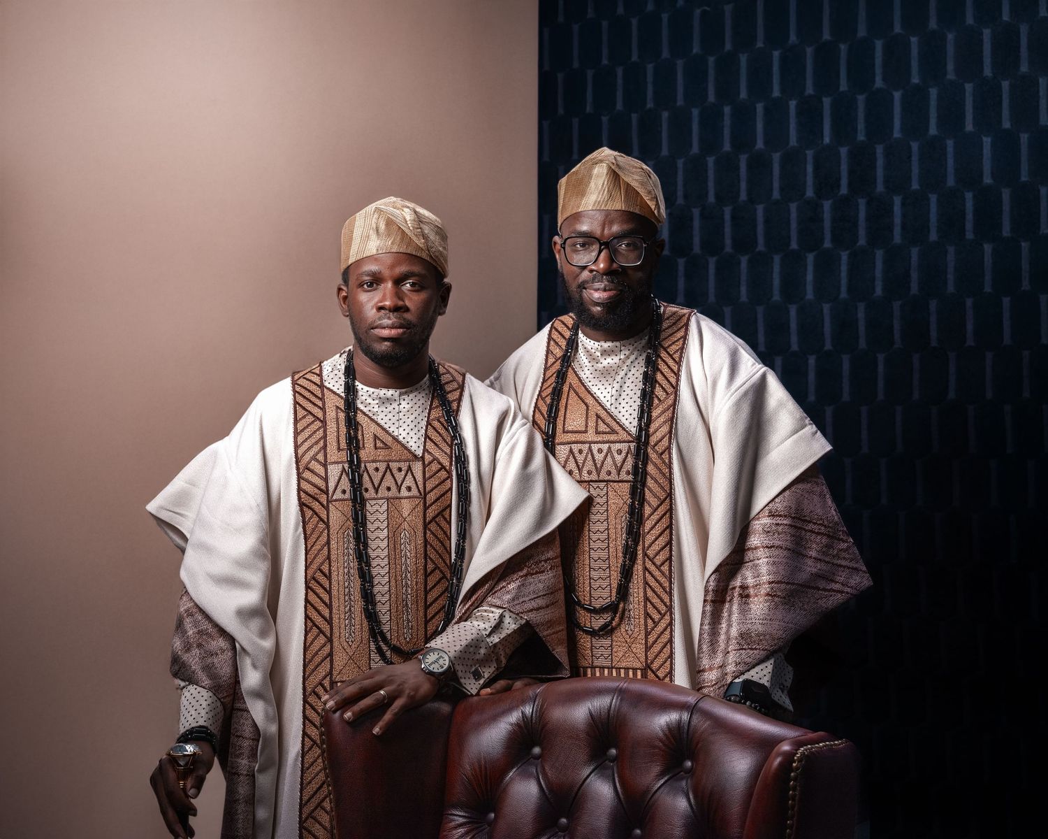 Two men in matching white and brown African Yoruba agbada outfits and kufi caps pose for a formal portrait.