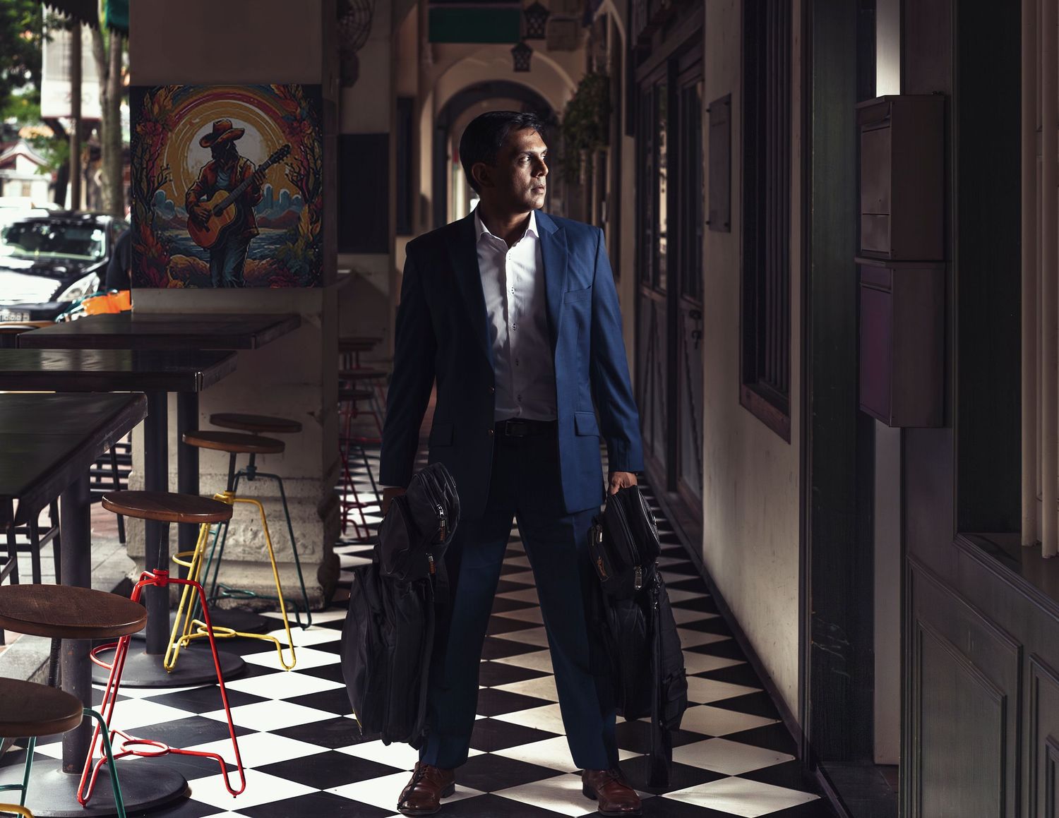 Man in blue suit carrying two black bags walks through a checkered floor corridor with colorful wall art.
