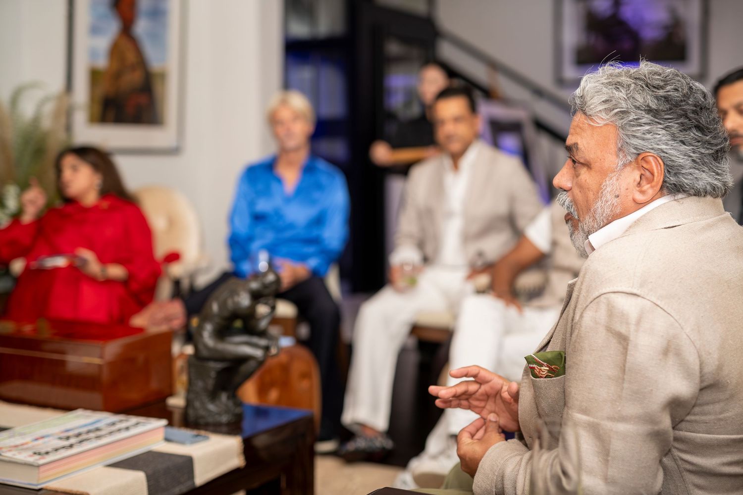 Man in linen suit speaks to seated audience during intimate indoor gathering or art event.