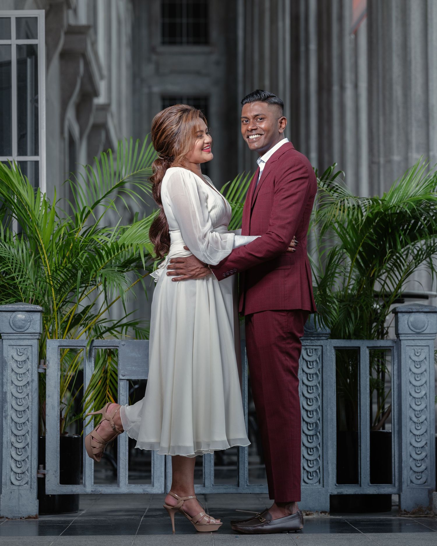 Couple in elegant attire, woman in white dress and man in burgundy suit, posing outdoors near tropical plants.