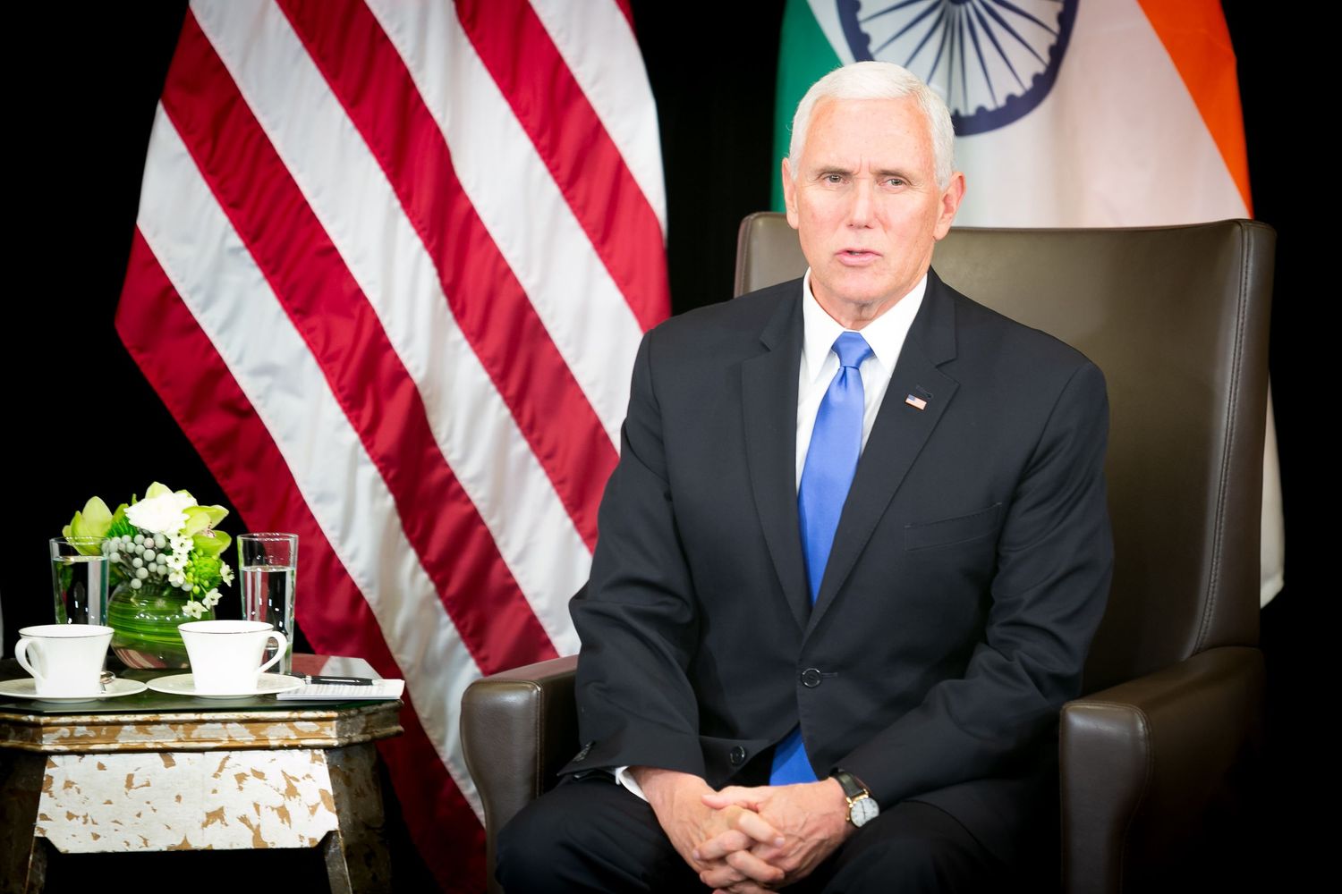 Man in suit sits before US and Indian flags in formal diplomatic meeting setting.