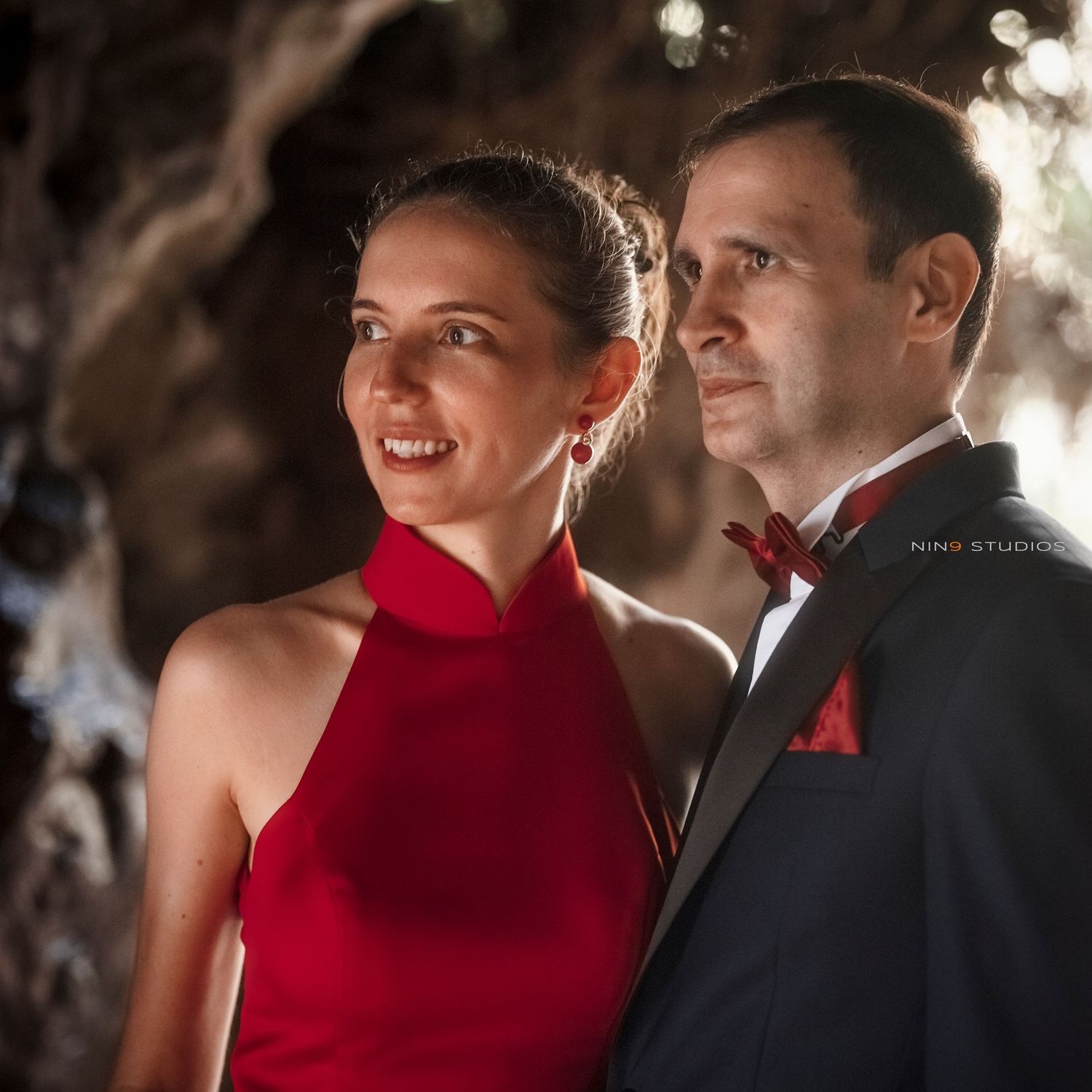 Elegant couple in formal attire, woman in red halter dress and man in black tuxedo with red bow tie, posing outdoors.